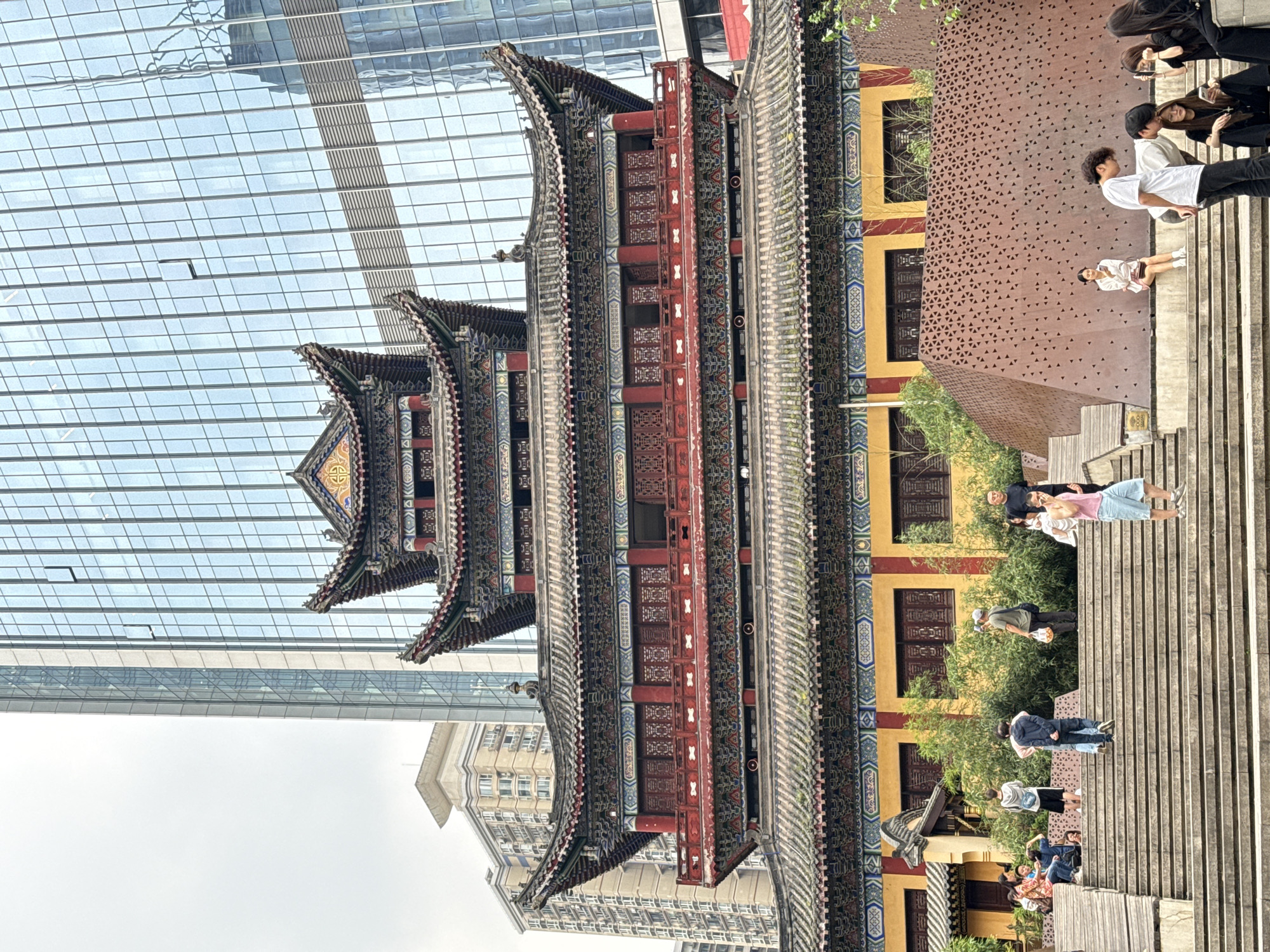 The image depicts a multi-tiered traditional Chinese pagoda in the Yuzhong District of China, juxtaposed with modern buildings in the background. The pagoda features intricate detailing on its eaves and vibrant colors on its facade. To the right of the pagoda is a series of wooden steps where several people are standing, sitting, or interacting. A building with a patterned brown facade and yellow-framed windows is visible adjacent to the steps. The sky is overcast, and the overall impression is one of a city blending traditional and contemporary architecture.
