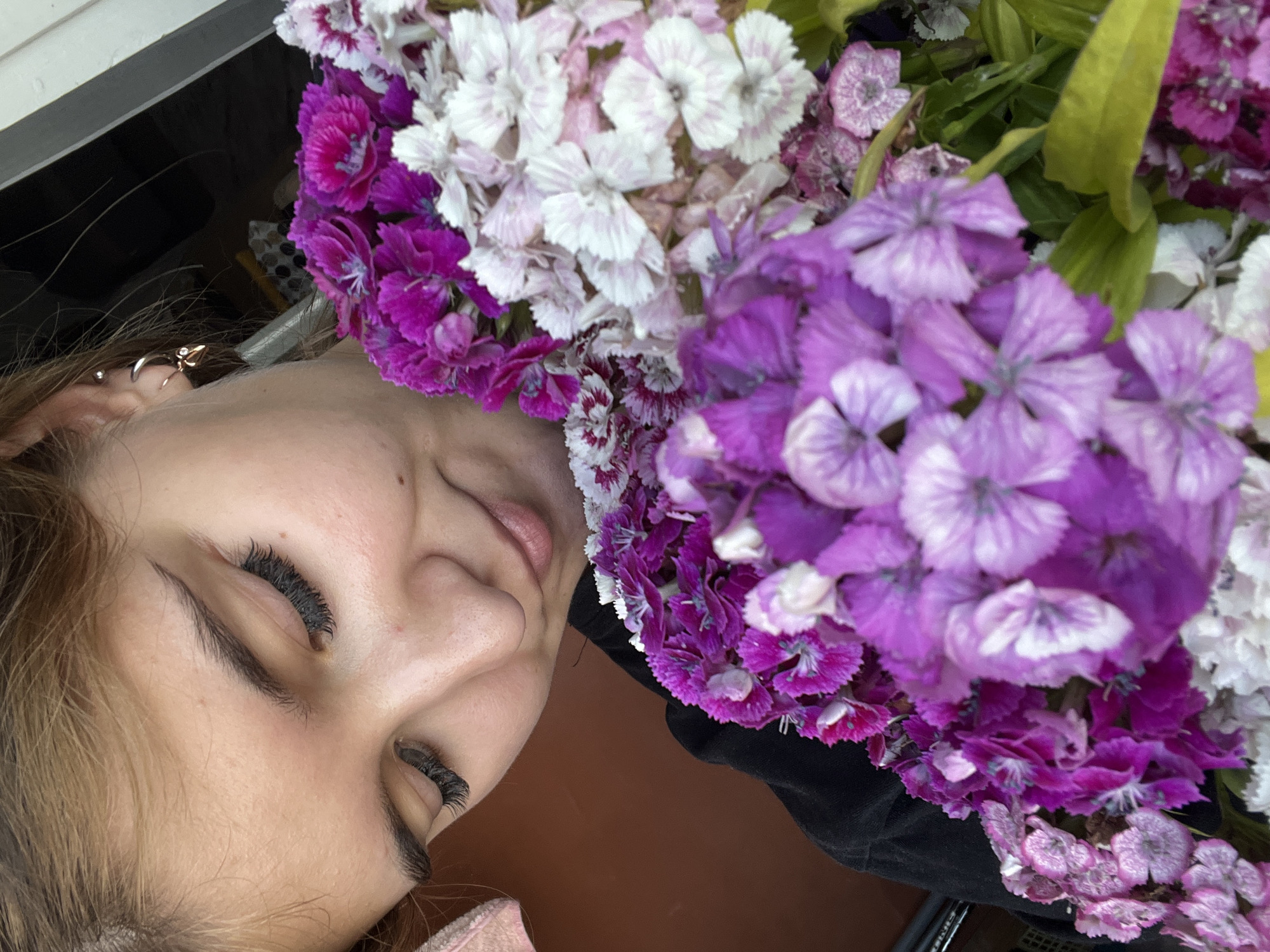 A close-up, outdoor, eye-level shot shows a young woman with light brown hair and dark eyelashes looking down at a bouquet of colorful flowers. She is wearing a black t-shirt and a silver earring with a gold accent in her left ear. The flowers are predominantly shades of purple and white, with some pink and magenta hues. The background is blurred, with hints of a brown wall and a dark, possibly outdoor, setting. The image appears to be taken in the city of Chaikovsky, Russia, judging by the context provided by the user.