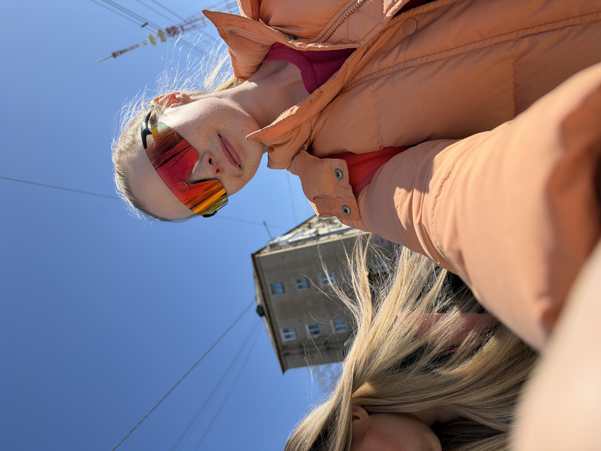 A low-angle selfie shows two women outside in Berezniki, Russia, with the sky and a building in the background. The first woman, in the foreground, is wearing a peach-colored winter jacket and red sunglasses. Freckles are visible on her face. Blond hair is visible in the lower portion of the frame, covering the face of the second person. Power lines are visible against the clear blue sky. The building in the background is a multi-story rectangular structure.