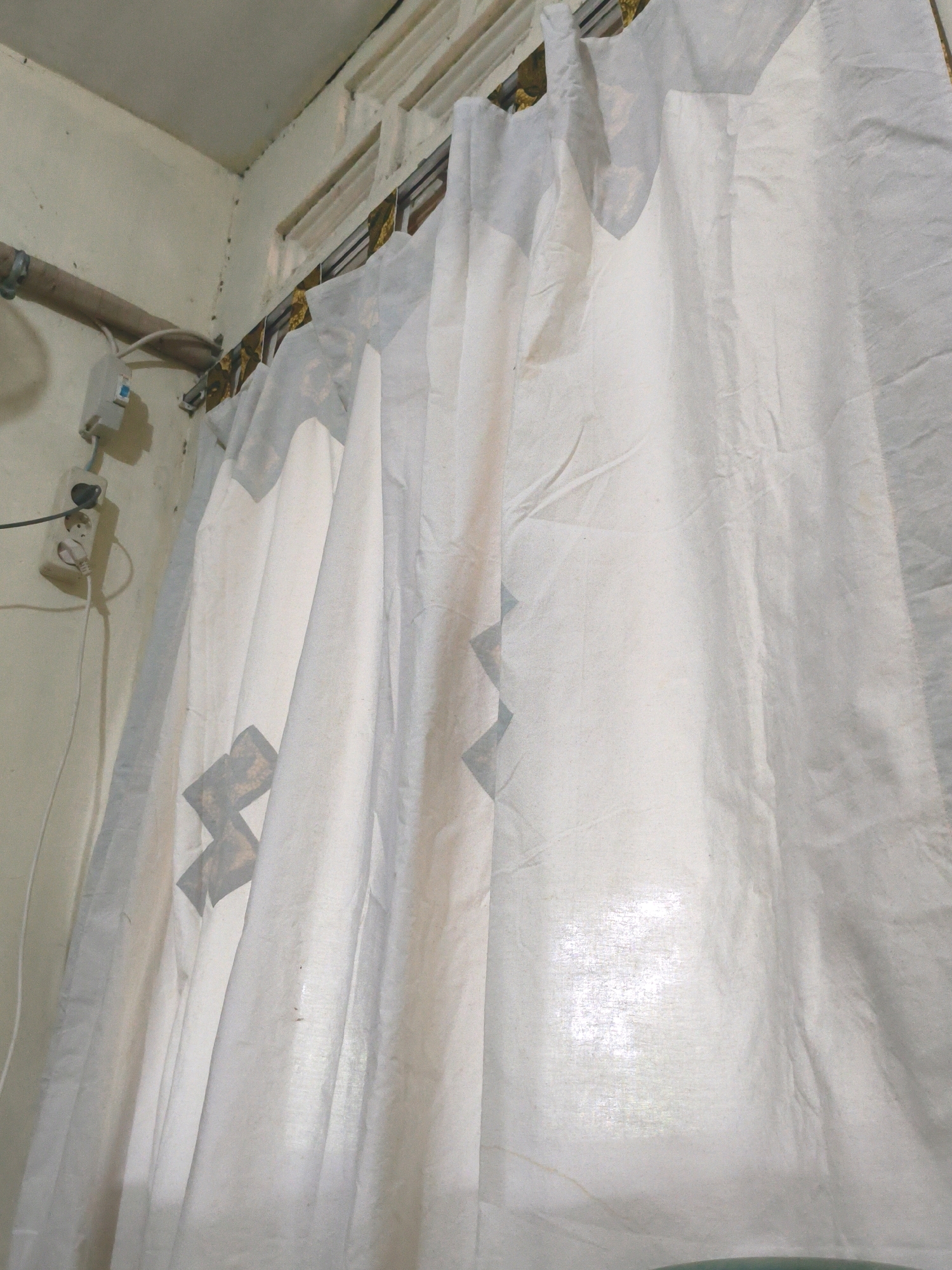 A close-up shot focuses on white curtains hanging from a curtain rod above a window in a room. The curtains are slightly sheer, allowing light to filter through from the window, creating a bright glow in the background. A subtle geometric pattern is visible on the curtains, with darker diamond shapes interspersed with lighter, possibly gold or tan, patterned elements. The curtain rod is mounted on the wall and appears to be made of metal. To the left of the curtains, a light-colored wall is visible, with a beige or off-white paint. A silver pipe is attached to the wall near the ceiling, and below it, a white electrical outlet with two sockets is mounted on the wall. A white electrical cord runs from the outlet. The image appears to be taken indoors, likely in a residential setting.