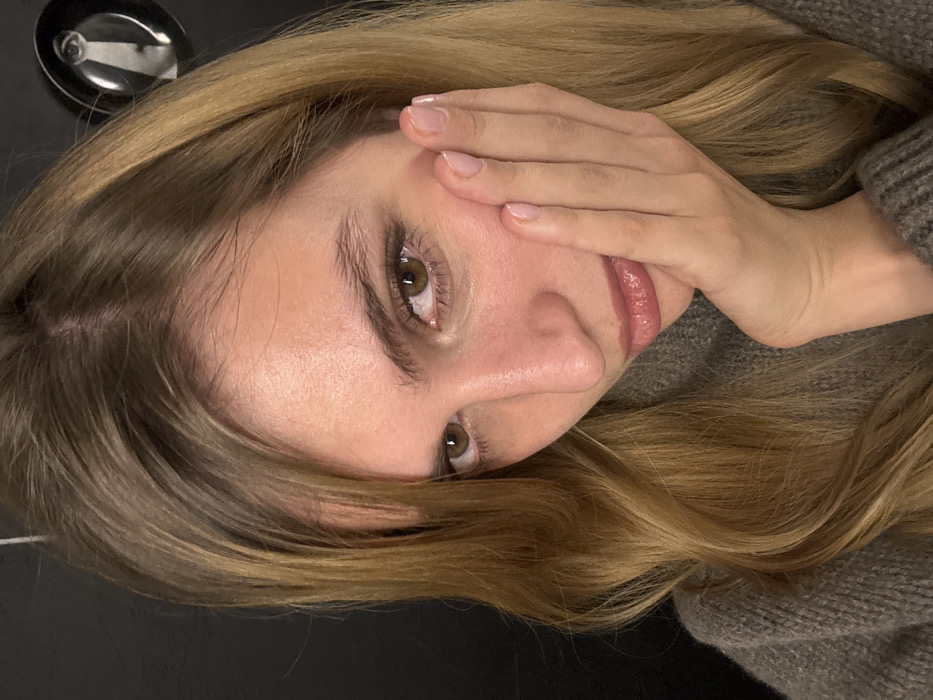 The image captures a close-up, tilted view of a person, likely female, lying on their side. The perspective is from slightly above the person's head, looking down onto their face.
The central subject is a person with fair skin and long, wavy, light brown or dark blonde hair with subtle lighter highlights. The hair is parted on the left side of their head and flows around their face and shoulder, some strands falling across their forehead and cheek.
The person's face is turned towards the viewer. Their eyes, which appear dark brown or hazel, are open and look directly forward. Both eyes are visible despite the person's right hand (viewer's left) covering the side of their face. Their eyebrows are well-defined and natural in color. The nose is prominent with a straight bridge. The lips are partially visible beneath the hand, with a soft, natural pink or mauve color, and appear slightly parted. The skin on the face appears smooth and clear, with a natural sheen.
The person's right hand is placed on the right side of their face, covering parts of the cheek and temple, and slightly obscuring the right side of the mouth. The nails are short, neat, and painted with a light, natural-looking polish. A small portion of a knitted garment, likely a sweater in a muted grey or light brown hue, is visible on the right side of the image, covering the person's shoulder or upper arm.
The background is largely dark and out of focus, possibly a plain surface like a table or floor. In the upper left corner of the image, a small, shiny metallic object, reflecting light, can be seen, resembling part of a spoon or a dish.
The lighting is soft and even, highlighting the features of the face and the texture of the hair. The overall impression is one of a relaxed or contemplative moment. No text is visible in the image.