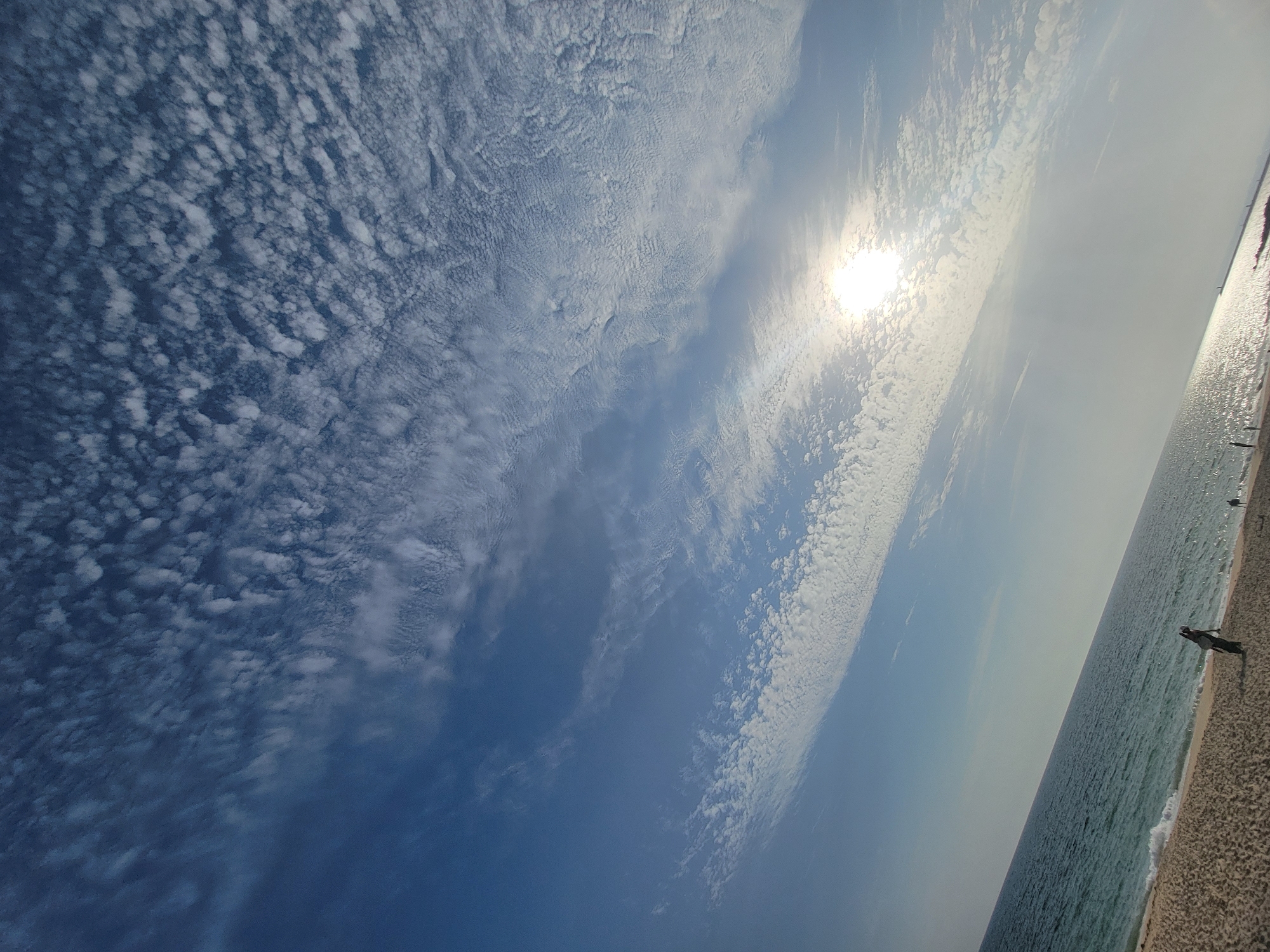 This media file captures a scene at Quarteira, Portugal, likely during the daytime, with a clear sky transitioning to a hazy horizon. The sun is bright and positioned in the upper right quadrant, casting a glare on the water and sand. The sky is filled with scattered white clouds that form textured patterns, particularly in the left half of the image. Below the sky, the ocean stretches to the horizon, its surface shimmering with sunlight and showing small waves breaking on the shore.
The foreground consists of a sandy beach, with the texture of the sand visible. A person is standing on the beach, appearing as a silhouetted figure facing away from the camera, looking out towards the ocean. Further down the beach, near the water's edge, another indistinct figure is visible. The overall impression is one of a calm day at the beach. The lighting suggests it could be mid-morning or late afternoon, with the sun at an angle. There is no visible text in the image.