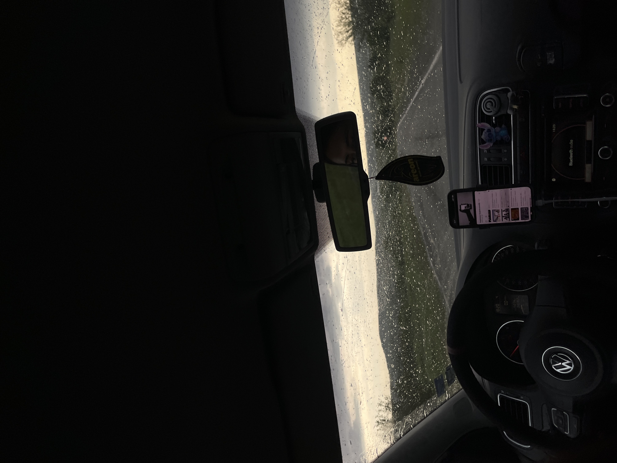 The interior of a Volkswagen vehicle is shown, with a focus on the dashboard, steering wheel, and windshield. Raindrops are visible on the windshield. A rearview mirror is attached to the windshield, reflecting a person. An air freshener hangs from the mirror. A smartphone in a mount displays a YouTube page on the dashboard. The radio is above the smartphone. The location is not evident and the city of Prilep, Bulgaria cannot be determined.