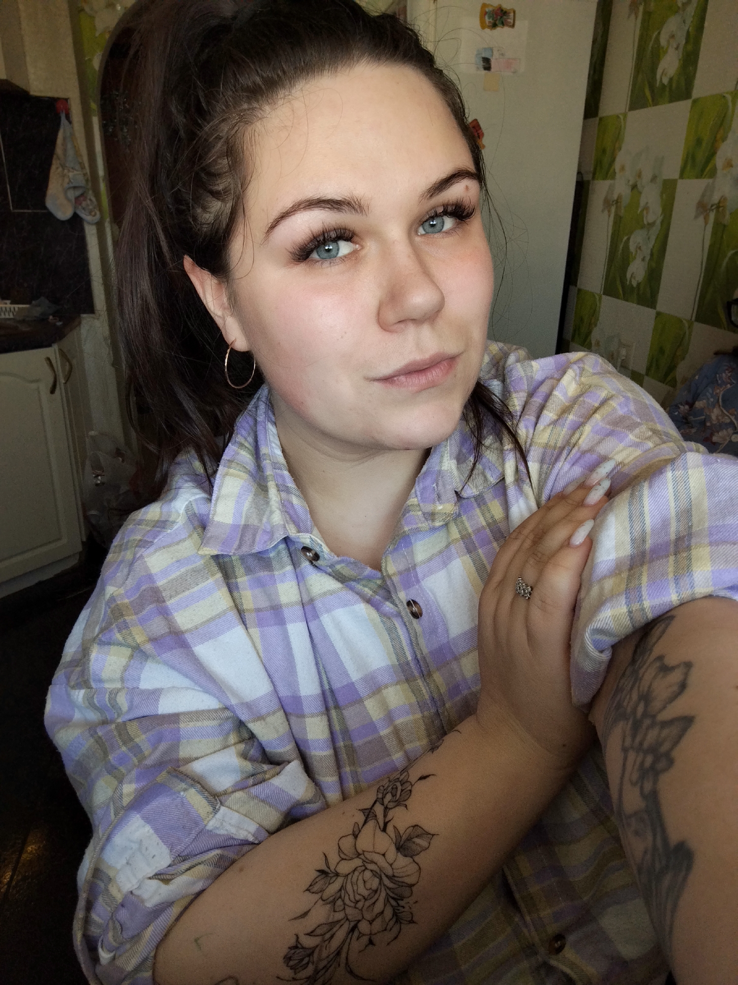 A medium-shot of a woman with fair skin and brown hair pulled back in a high ponytail. She has striking blue eyes and is wearing large, false eyelashes. She has a small gold hoop earring in her left ear and is wearing a light purple and white plaid button-down shirt. Her right arm is bent and held across her chest, and she has long, light-colored nails and a ring on her right ring finger. Two visible tattoos are on her arms, both floral designs in black ink.
The background is a combination of domestic settings. To the left is a white kitchen cabinet, and to the right, a wall with light-colored wallpaper patterned with white and green flowers. The scene is dimly lit, suggesting indoor lighting. There is no visible indication that this scene is located in Chaikovsky, Russia.