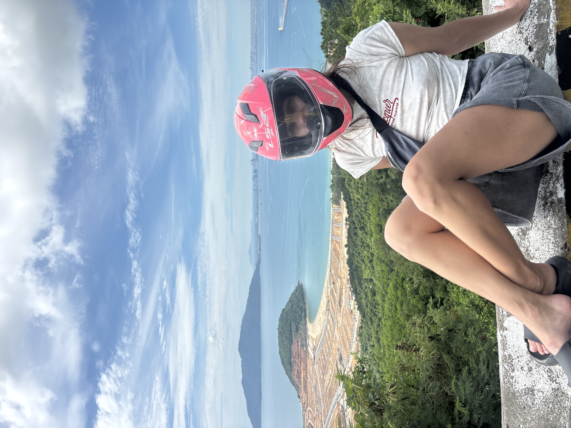 A young woman wearing a pink motorcycle helmet sits on a concrete ledge, looking at the camera. She has on a white t-shirt with text and gray denim shorts. She is leaning back with her legs crossed and her left foot resting on a black sandal. A dark gray bag is slung across her chest. The background shows a panoramic view of the city of Đà Nẵng, Vietnam. In the middle ground, a peninsula with a beach and construction sites is visible, with the sea stretching out beyond it. Further in the distance, mountains and a cityscape can be seen under a partly cloudy blue sky.