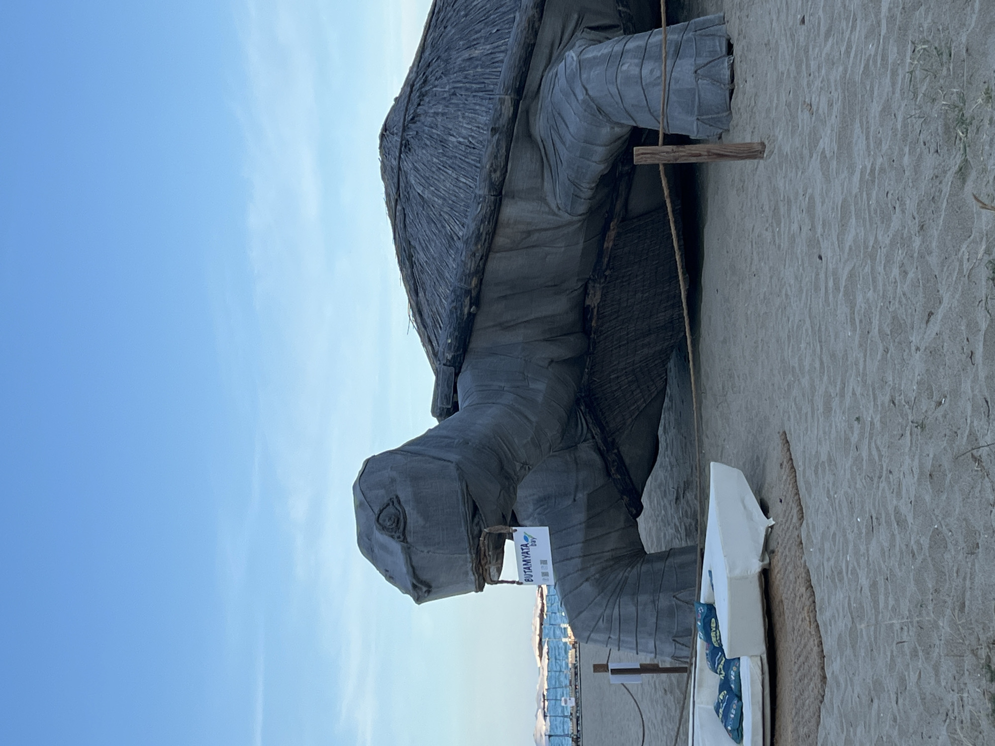 An outdoor daytime shot shows a large, grey, stylized tortoise sculpture on a sandy beach. The sculpture is made of textured material, possibly fabric or canvas, stretched over a frame. Its shell is thatched with straw or similar material. A sign hangs from the tortoise's mouth, reading "BUTAMYATA bay" and featuring a QR code. To the left of the sculpture, a white sofa with decorative blue pillows rests on a woven mat. In the background, there is a body of water, beach umbrellas, and a clear blue sky. The scene appears to be in a resort or beach area.