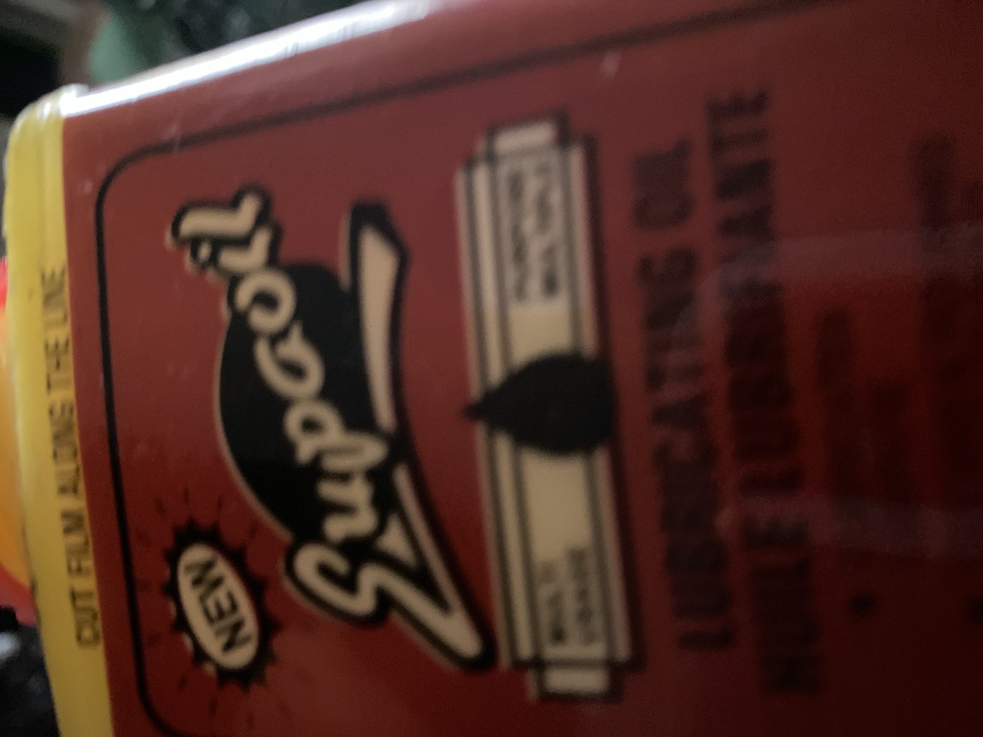 The image displays a close-up of a red metal can, likely a lubricant product. Prominently featured is the brand name "3-IN-ONE" in stylized white lettering with a black outline, and the word "Supaoil" below it in a similar style. Above "Supaoil", there is a black oval shape with "NEW" written in white lettering inside. Below the main branding, there are two horizontal white bars with text that is partially obscured and out of focus. The top of the can has a yellow band with the text "CUT FILM ALONG THE LINE" printed in black. The bottom of the can shows the words "LUBRICATING OIL" and "HUILE LUBRIFIANTE" in black lettering. The overall condition of the can appears worn, with some smudges and a grainy texture to the image. The background is dark and out of focus, making it difficult to discern any environmental details. There are no people or any identifiable elements of the city of Jalingo, Nigeria, present in the image.