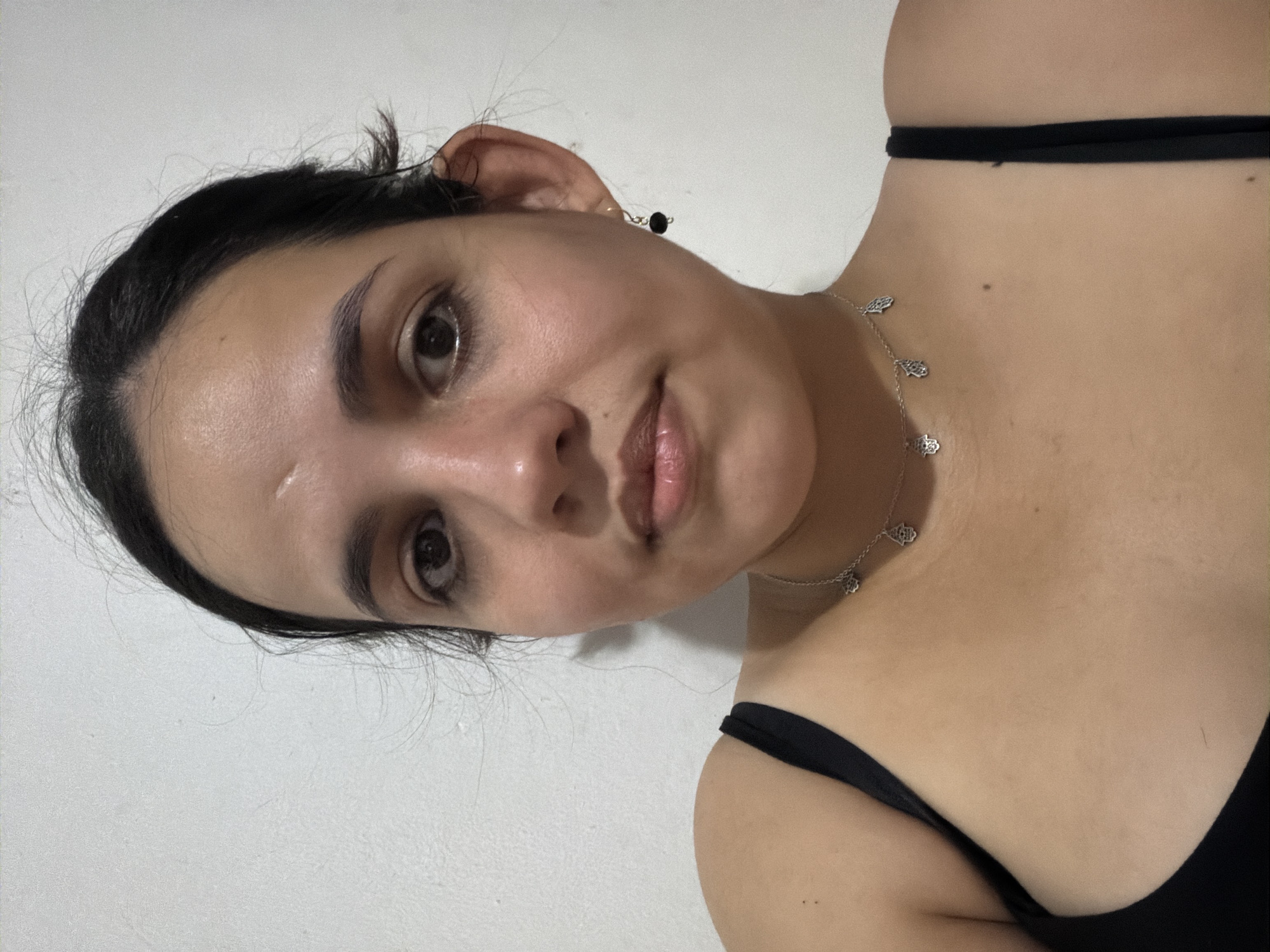 A woman with dark hair and brown eyes is facing the camera. She is wearing a black top and a silver necklace with hand-shaped pendants. She has a dark earring on her left ear. The background is a plain white wall. There is no information in the image to determine her location in Ciudad Guayana, Venezuela.