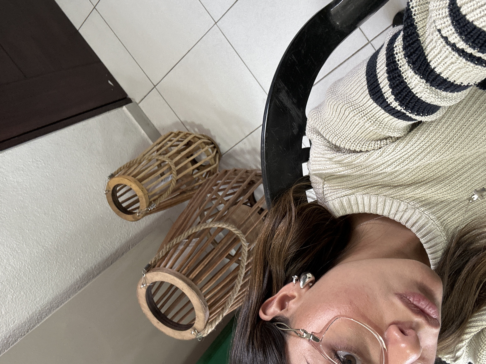 A medium-shot, high-angle photograph displays an individual seated in a black chair, wearing a cream-colored sweater with black stripes. The person is looking to the right, wearing glasses and multiple earrings. Beside them, on a white-tiled floor, sit two decorative, cylindrical wooden lanterns. Adjacent to the tile floor is a darker brown surface and white wall, potentially a door or cabinet. The location appears to be indoors, possibly within a residence. The exact city and specific location are unknown.