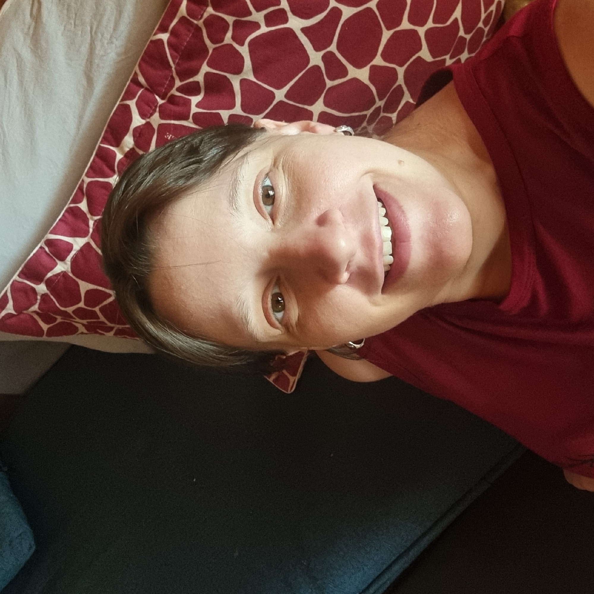 A woman with brown hair and brown eyes smiles at the camera. She is wearing a maroon shirt and silver hoop earrings. She is lying down next to a pillow with a giraffe print pattern. The background is dark and out of focus, suggesting the photo was taken indoors. There is no information available about the location in the city of Dunnae-myeon, South Korea.