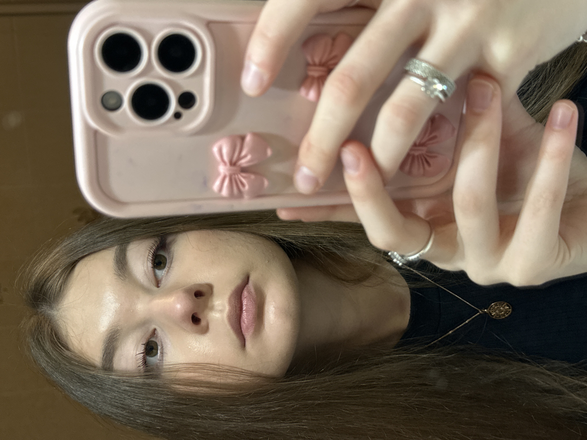 The image depicts a close-up selfie of a young woman, taken in what appears to be a reflective surface, possibly a mirror. Her head is tilted, indicating the photograph was taken with the phone held horizontally and then rotated. The woman's face is prominent, featuring brown eyes looking directly at the camera, light brown hair framing her face, and a neutral expression with slightly parted, natural pink lips. She appears to be wearing light makeup, including mascara.
She is holding a smartphone in her hands, which are visible in the upper part of the frame. The phone is encased in a light pink protective cover, adorned with three distinct, raised bow-shaped embellishments of a similar pink hue. The phone's camera module area is visible, showing cutouts for what appears to be three large lenses, a smaller sensor, and a flash, characteristic of modern smartphones like recent iPhone Pro models.
On her left ring finger, she wears a silver-toned ring with multiple small stones and a larger central stone. A simpler silver band is visible on her right middle finger. Around her neck, a delicate silver-toned necklace with a small, circular pendant featuring an intricate, possibly sun-like, design is visible.
The background is uniformly blurred and appears to be a plain, light brown wall or surface, suggesting an indoor setting. There are no specific visual cues within the image itself to indicate the provided location context of Kazan, Russia. No visible text is present in the image.