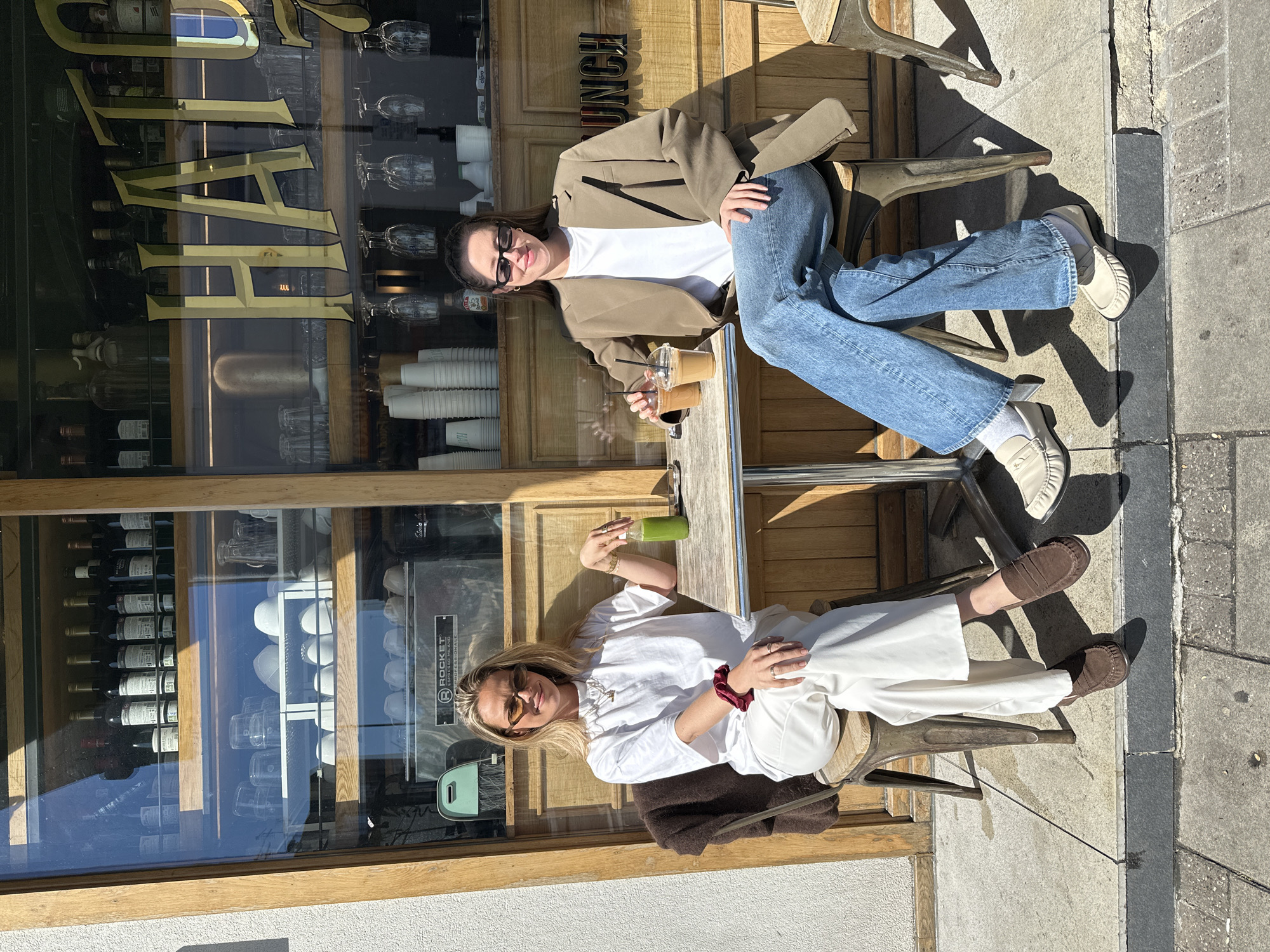 Two young women are seated at a small table outside a restaurant. The woman on the right wears a tan blazer, white t-shirt, blue jeans, and off-white loafers. She has sunglasses on her head and holds two drinks. The woman on the left wears a white top and pants, brown loafers, and sunglasses. She also has a drink on the table. The restaurant has a large window displaying wine bottles and cups. "Lunch" is written in gold lettering above the window. The setting appears to be an outdoor cafe. Based on the style of architecture, including the facade of the building in which the cafe is located, the location could be London, United Kingdom.