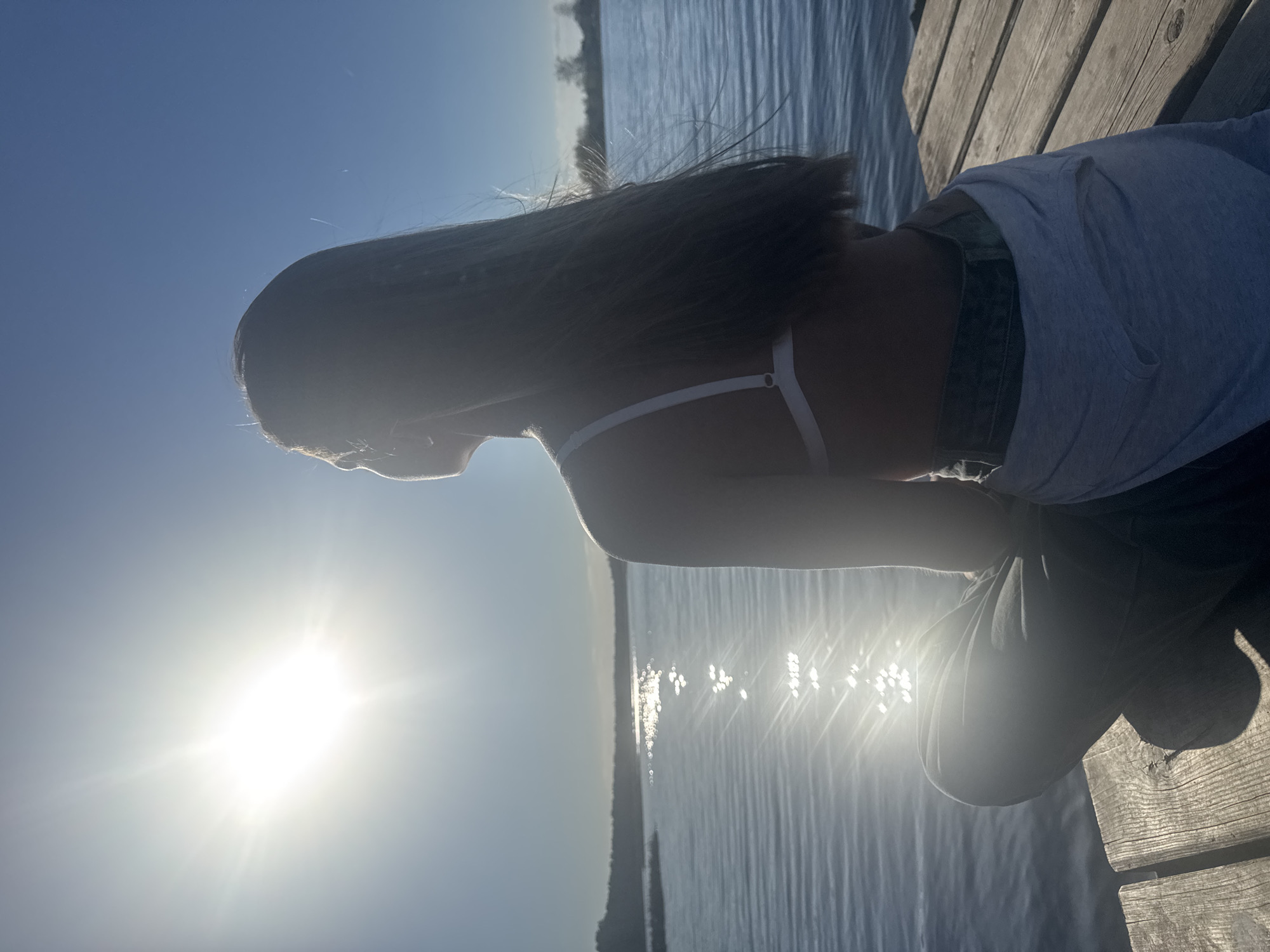 A young person with long hair is sitting on a wooden dock, facing away from the camera and toward a body of water. The sun is prominently positioned in the sky, creating a bright glare. The person is wearing a light-colored top and dark pants. The location appears to be near a body of water, with trees visible in the distance. The exact location within the city of Костенево, Russia is not identifiable based solely on the image.