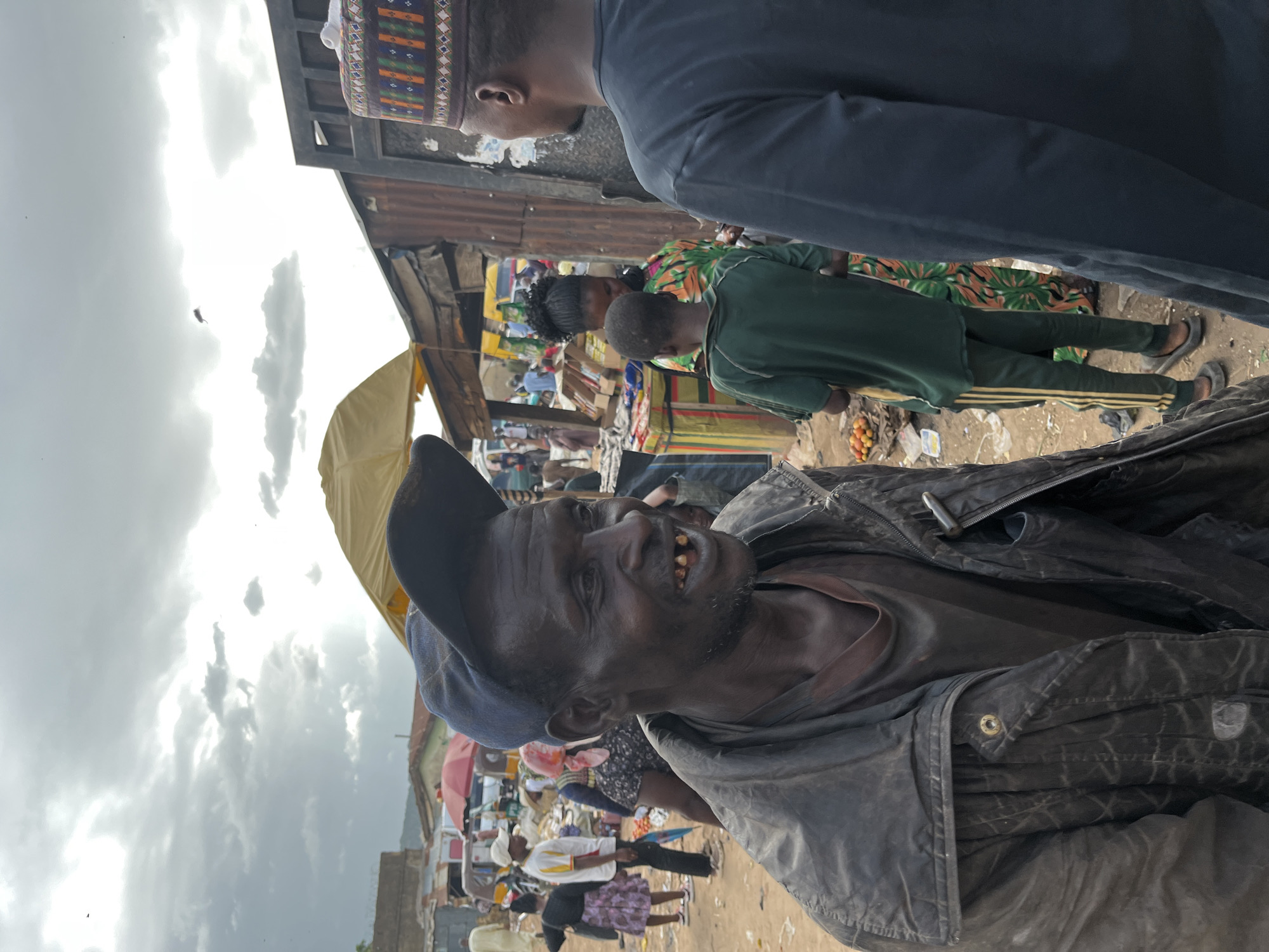 The media file depicts a lively outdoor market scene in Rafin Par, Nigeria. The foreground is dominated by a close-up of an elderly man with dark skin, wearing a dark jacket and a blue baseball cap. He has a weathered face, a visible beard, and an open mouth revealing several missing teeth, suggesting a candid and perhaps humorous moment captured. His expression seems to be one of amusement or perhaps weariness, looking towards the viewer.
Behind him, the market bustles with activity. People are seen milling about, engaged in shopping or interacting. A woman in a vibrantly patterned green, orange, and white dress stands out, alongside a young boy in a dark green shirt and pants. Further in the background, stalls are visible with various goods, suggesting a typical market environment. The presence of umbrellas and awnings indicates an attempt to shield from the sun.
The time of day appears to be daytime, with bright sky and clouds visible. The weather seems to be overcast or partly cloudy. The overall setting is a crowded marketplace, characteristic of many towns and cities in Nigeria, with corrugated metal structures and dirt ground visible. The image is taken at a slight angle, giving it a dynamic feel. There is no visible text in the image.