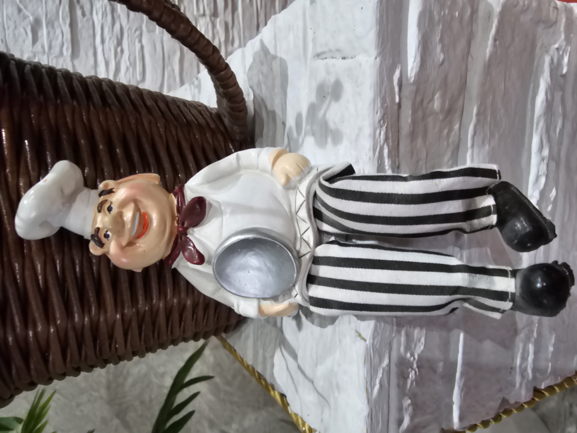 A ceramic figurine of a jolly chef sits on a textured white surface. The chef wears a white chef's hat, a white shirt with a maroon bow tie, and black and white striped pants. In its hands, the chef holds a silver ladle. The figurine is positioned in front of a large, dark brown woven basket. The background consists of a light-colored textured wall. The scene appears to be an indoor decorative display, potentially in a restaurant or shop, and there is no indication of its location in Kalinovka, Russia.