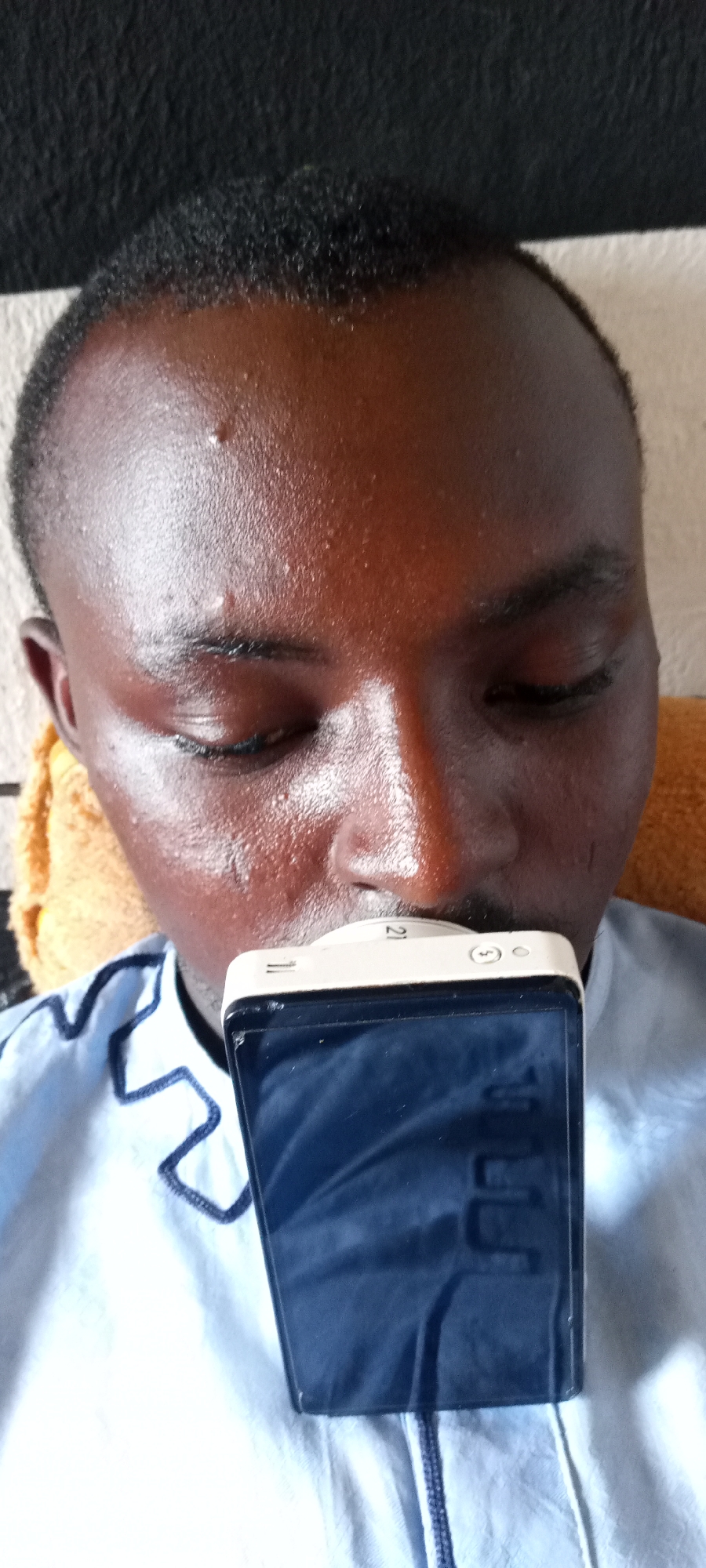 A close-up shot features a dark-skinned man with his eyes closed and a mobile phone held against his mouth. The man has short, dark hair and a slightly sweaty face. He is wearing a light blue shirt with a dark blue zig-zag pattern around the collar. The phone is primarily black with a white top, and the screen reflects a blurred image. The background includes a textured surface above the man's head and what appears to be a yellow cloth or pillow behind his head. The location is speculated to be in Jos, Nigeria.