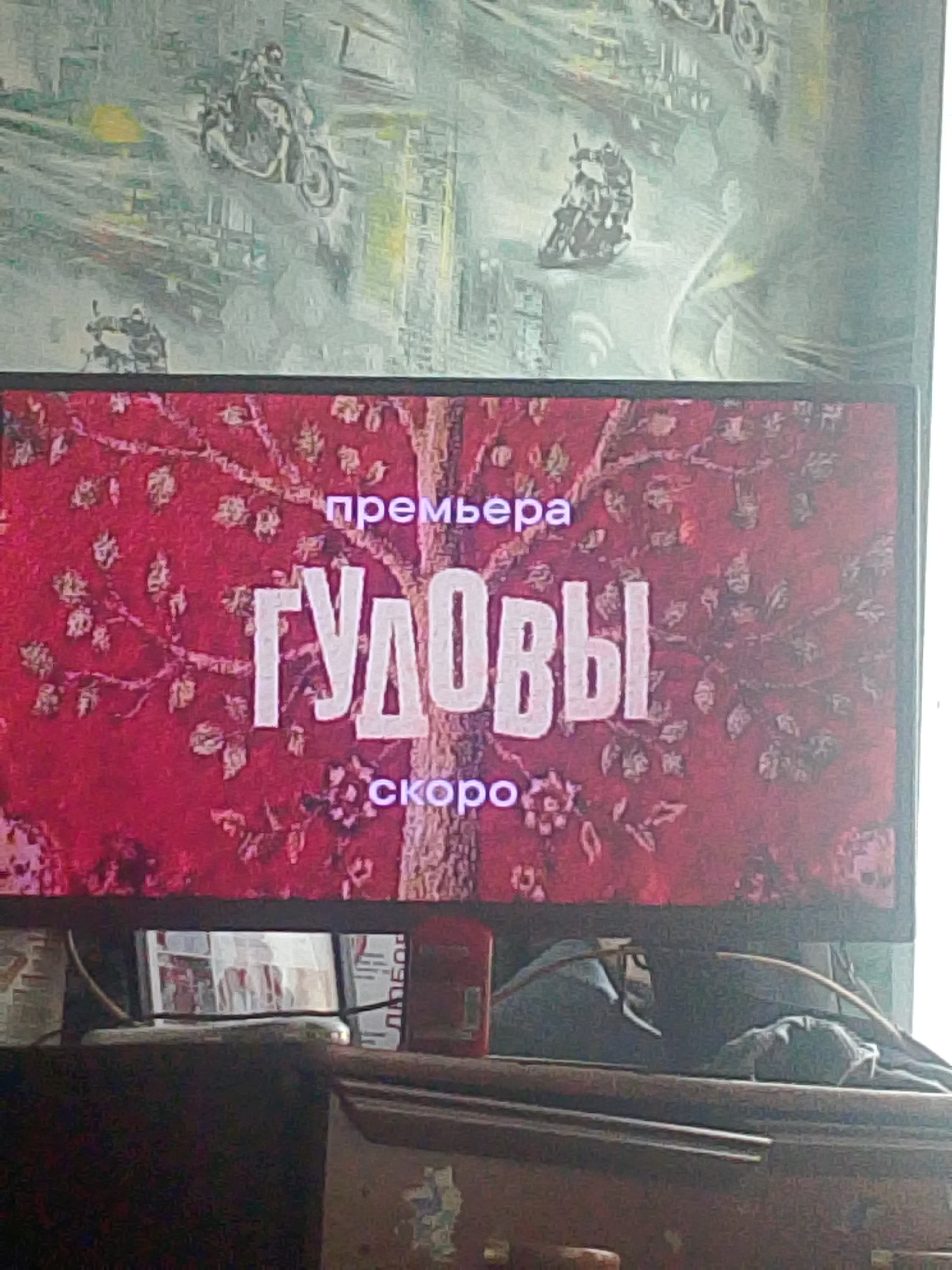 The image displays a television screen showing a preview for an event with Russian text. The screen is mounted above a wooden dresser or cabinet. The text "ПРЕМЬЕРА" (PREMIERE) is visible at the top, followed by the larger text "ГУДОВЫ" (GUDOVY). Below this, the word "СКОРО" (SOON) is displayed. The background of the screen features a red textured pattern with tree branches and leaves. The overall scene is indoors. There is no discernible location in the city of Asino, Russia, present in the image.