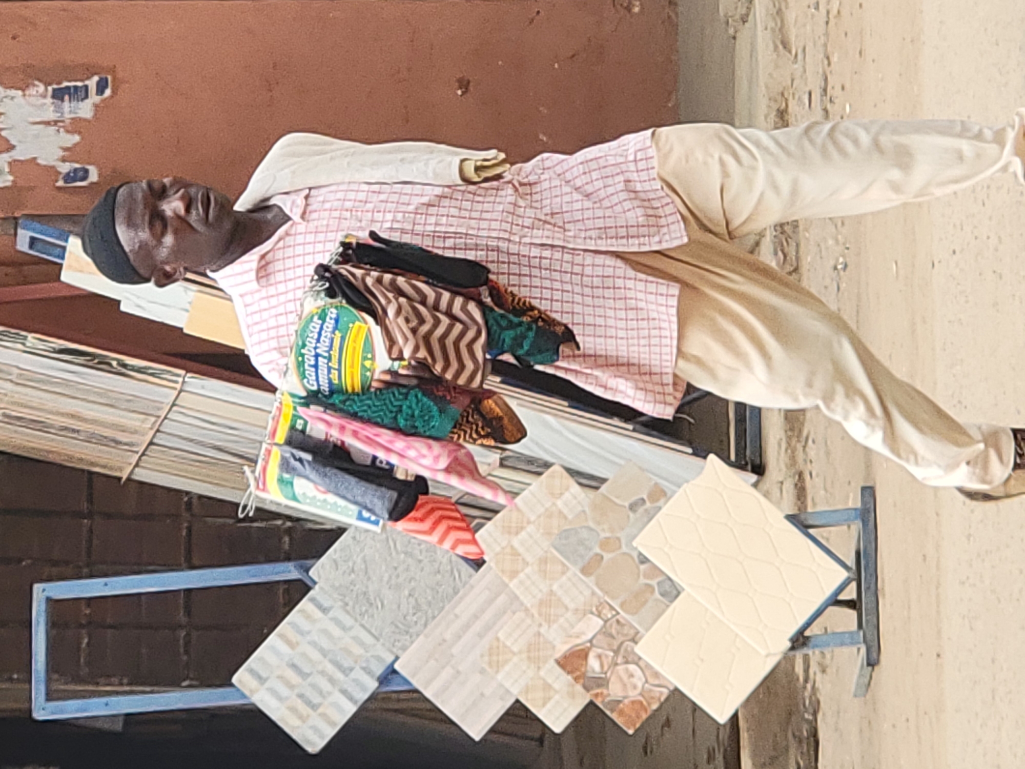In Maiburiji, Nigeria, an individual wearing a black cap, a red and white checked shirt, and light beige pants, lies on an assortment of objects, including tiles of different patterns and a package labeled "Garabasar Jami'un Nasara da Indomie". The objects are arranged on a small blue metal stand. The background features a brown wall with peeling paint and an outdoor, possibly street-side, environment.