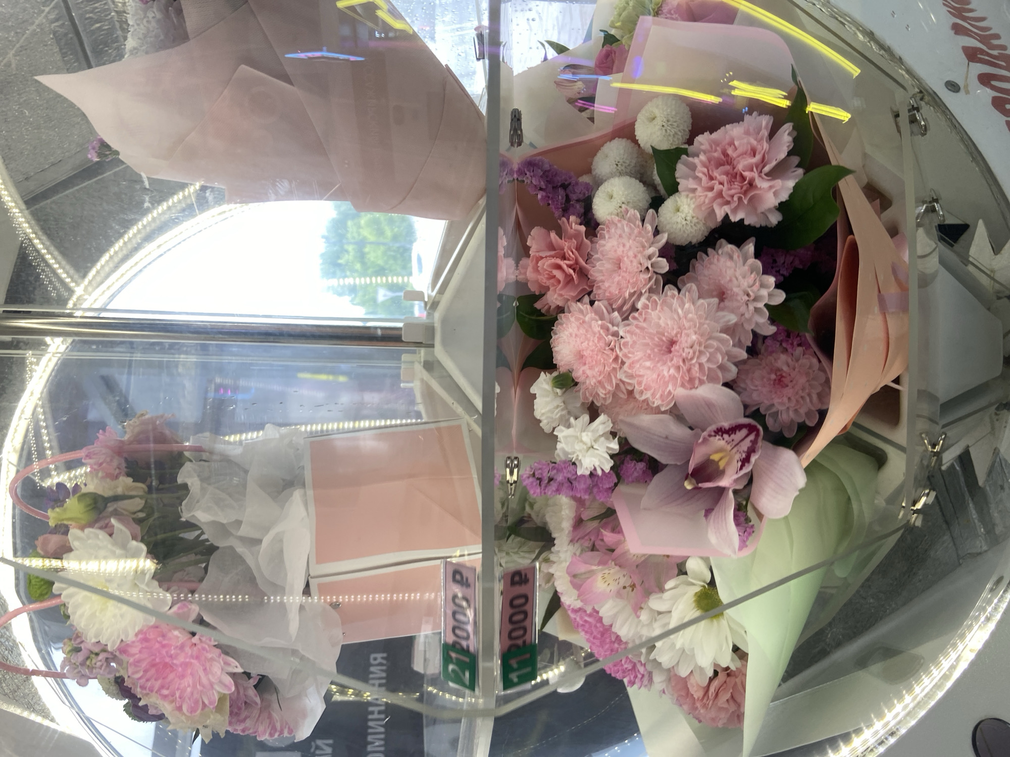The image displays a close-up of floral arrangements displayed in a glass enclosure, likely a vending machine. The flowers are predominantly in shades of pink, white, and purple. Two prominent price tags are visible within the enclosure, one with the number "21" and the other with "11", both followed by "2000 P", indicating a price of 2000 rubles. The background shows a blurred view of trees and a street, suggesting an outdoor urban setting. Text visible on the enclosure includes what appears to be Russian lettering. The overall impression is a commercial display of flower bouquets for sale. The user has requested the location to be Ufa, Russia.