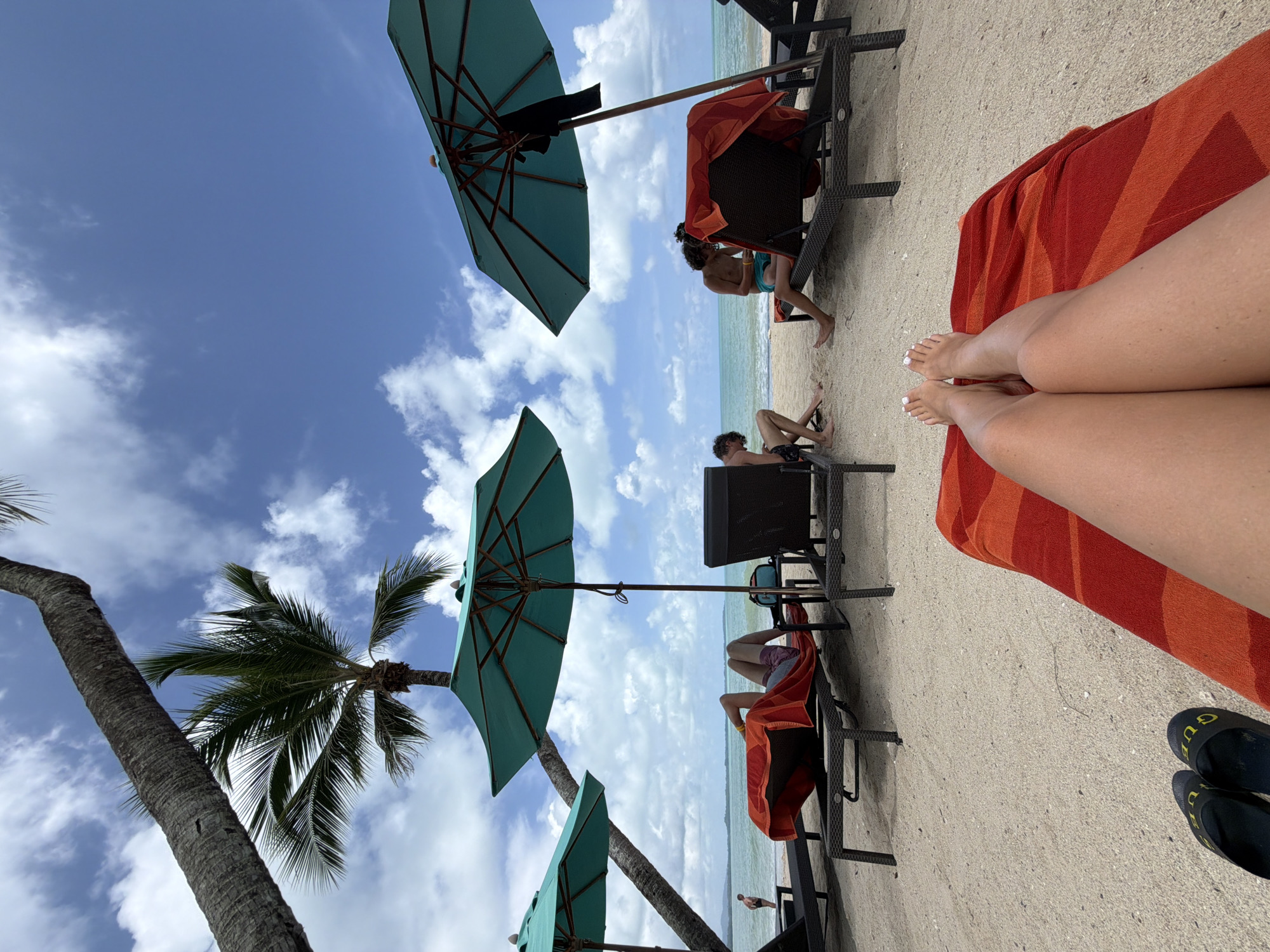 The image depicts a beach scene in Bang Muang, Thailand. In the foreground, a person's legs, covered by a red and orange striped towel, are visible, resting on a lounge chair. A pair of dark sandals with yellow lettering are placed next to the chair on the sand. In the middle ground, several lounge chairs are set up on the sandy beach, some occupied by people. Two teal-colored beach umbrellas provide shade. One person is lying on a lounge chair under an umbrella, covered by a red towel, with their legs visible. Another individual is sitting on a lounge chair, looking down at something in their hands. A third person is partially visible sitting on a lounge chair. In the background, the ocean stretches out to the horizon under a bright blue sky with scattered clouds. Palm trees frame the scene, with their fronds casting shadows. A single person can be seen walking along the shoreline in the distance.