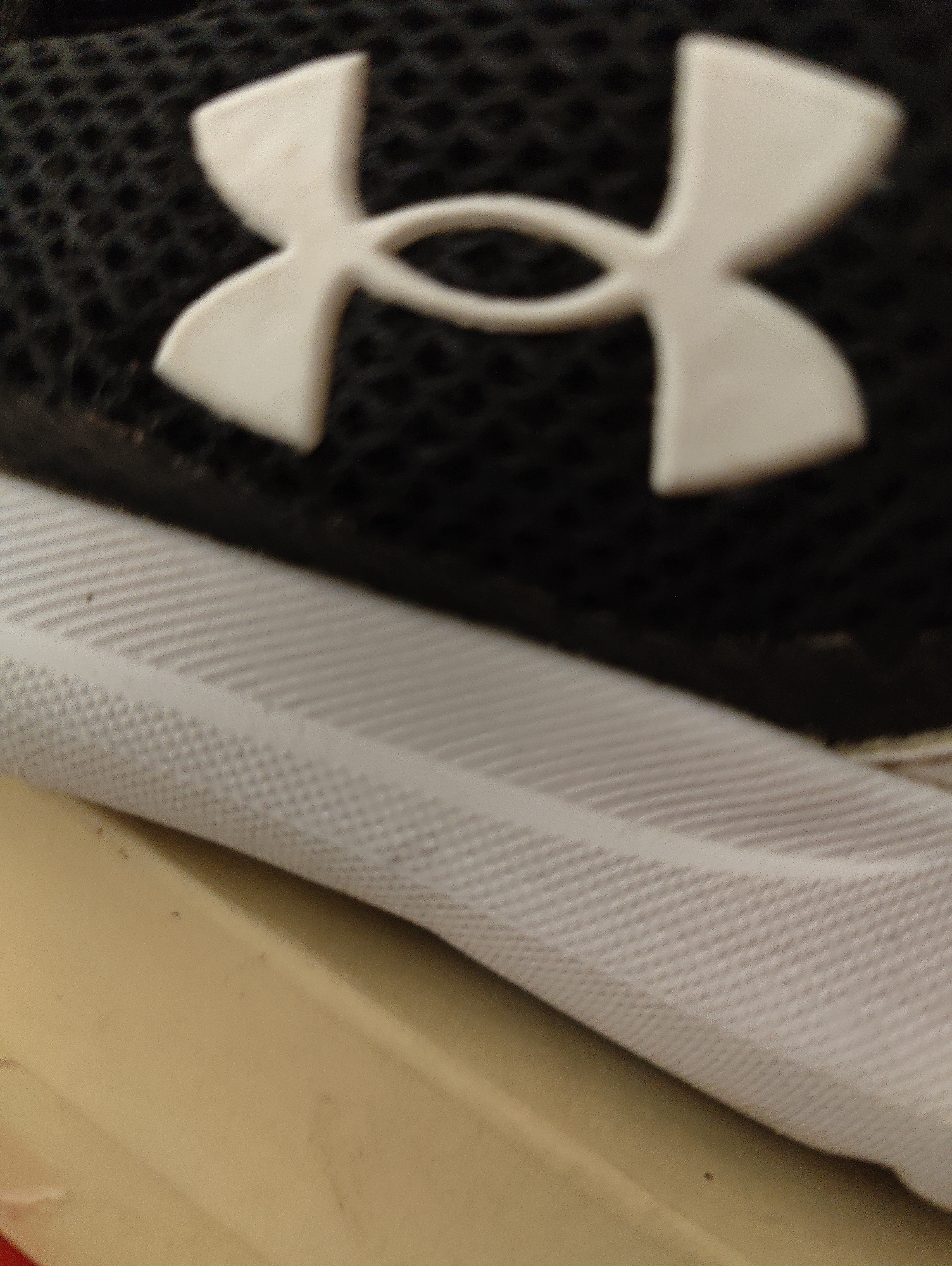 A close-up, angled shot shows a portion of a black athletic shoe with a white Under Armour logo. Below the shoe is a textured white material. The background is a light brown, out-of-focus surface. The scene is indoors, with no visible city context.