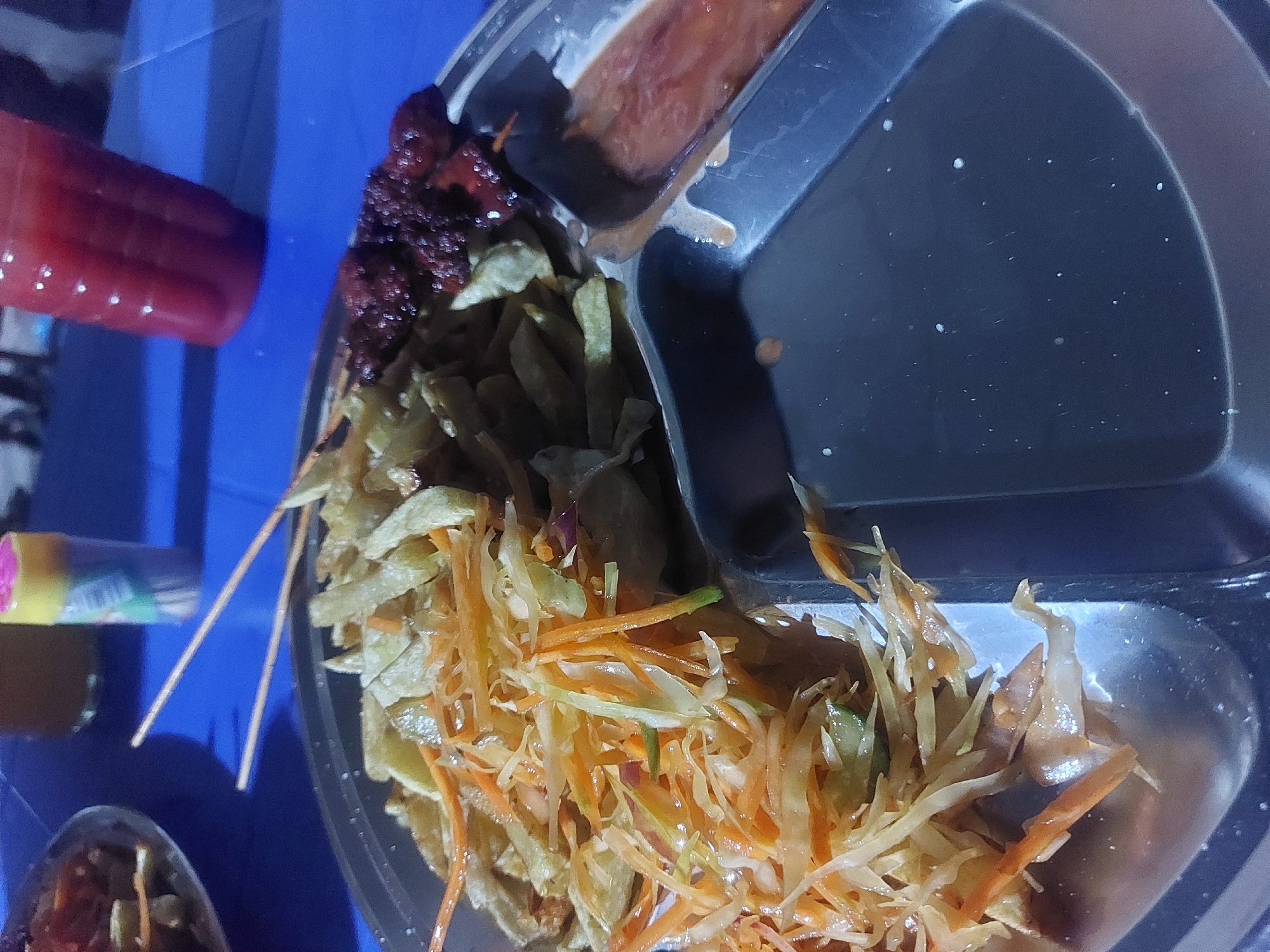 A three-compartment tray of food sits on a blue plastic surface. One compartment holds a dark red sauce; another compartment is empty; the third contains a shredded mixture of vegetables, including carrots. Beside the tray are skewers with dark red meat and fried chips. In the background, there is a red drink, a yellow tube, and a small metal container. Based on the food and presentation style, it is likely to be a street food meal from Dar es Salaam, Tanzania.