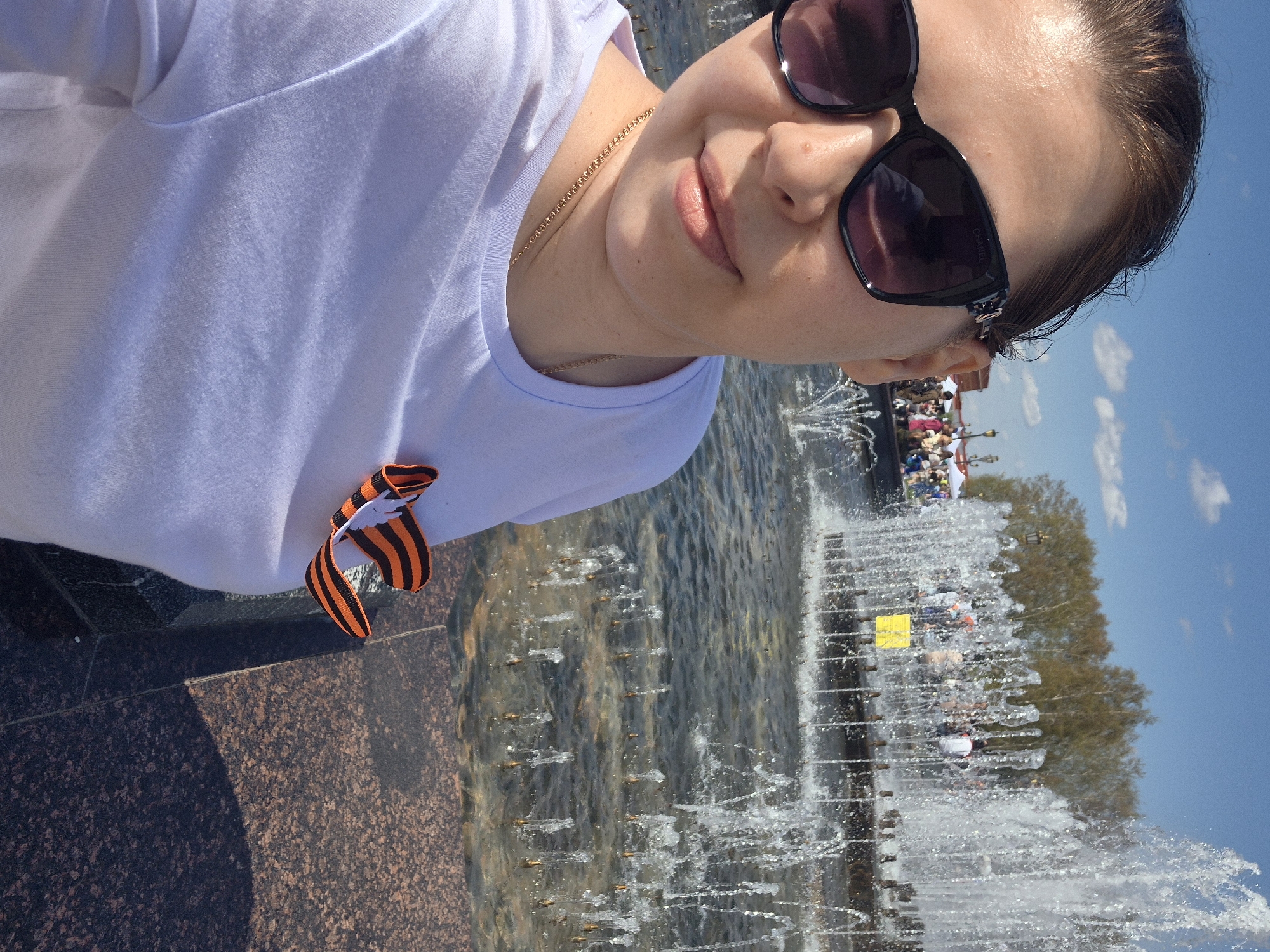A woman with brown hair and sunglasses is taking a selfie near a fountain. She is wearing a white t-shirt and a black and orange ribbon. In the background, there are trees and people, suggesting a public space. The location is Tobolsk, Russia, as evidenced by the prevalent local celebrations and the architectural style of the structures in the background, aligning with common landmarks found in this area. The sunlit setting suggests a clear day, and the fountain provides a dynamic element to the scene.