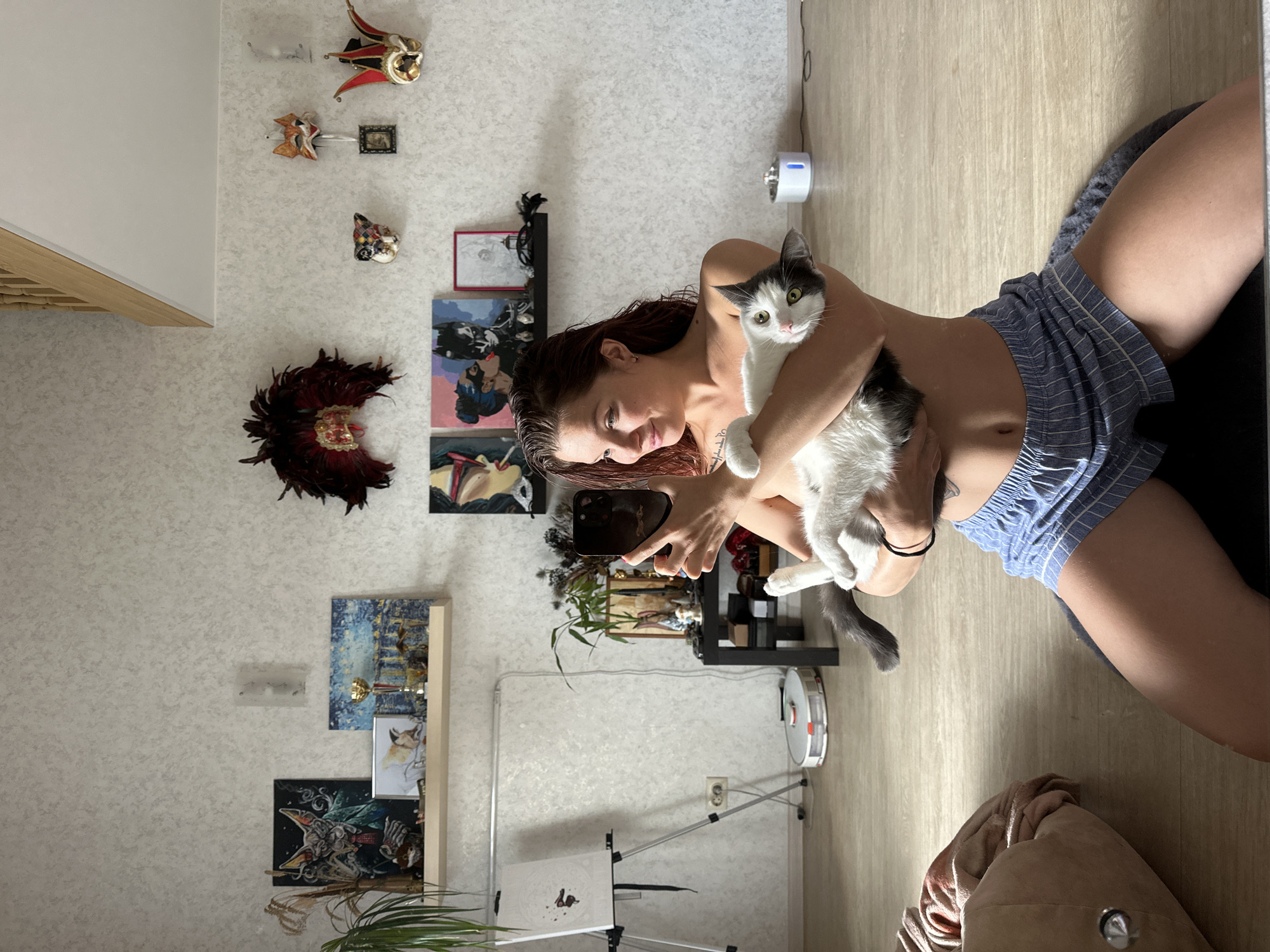 A young woman is kneeling on the floor, holding a black and white cat. She is looking at her phone, which she holds in her left hand, and appears to be taking a selfie. She is wearing blue and white striped shorts. The cat is looking at the camera. In the background, there are paintings on the wall, a shelf with various items, and a robot vacuum cleaner on the floor. The room has light-colored walls and wooden flooring. There is a window and a wooden staircase in the background. The scene appears to be an indoor setting, likely a living room or bedroom, possibly in an apartment. There is no explicit information in the image that can definitively place the location in Vladimir, Russia.