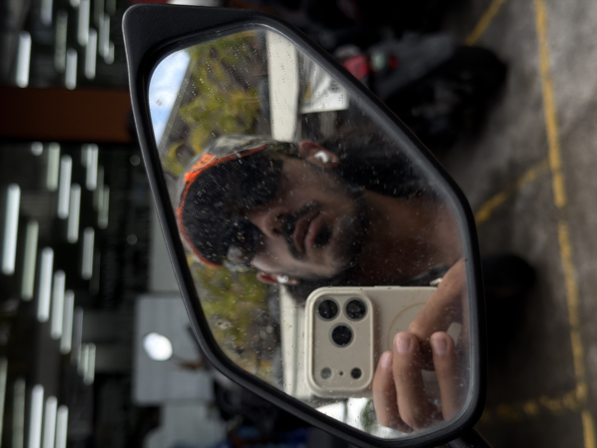 A diagonal, close-up shot features a black rearview mirror, likely from a motorcycle or scooter, capturing a reflection-based selfie in North Kuta, Indonesia. Within the mirror's surface, a person's reflection is visible, wearing dark sunglasses and a dark head covering with an orange band. An earbud is visible in their right ear. The person is holding a light-colored smartphone with a distinctive triple-lens camera system and a flash, visible in the lower right portion of the mirror, held by their right hand.
The mirror's surface is speckled with numerous small water droplets, suggesting recent rain or condensation. The reflection inside the mirror includes a cloudy sky, green foliage, and faint yellow lines on a dark, possibly asphalt, surface, indicating an outdoor setting. Outside the mirror's frame, the background is softly blurred, showing vertical white streaks, possibly lights or architectural elements, and a horizontal brown section, suggesting a partially covered or indoor area in the immediate surroundings. The scene takes place during daylight. No visible text is present.