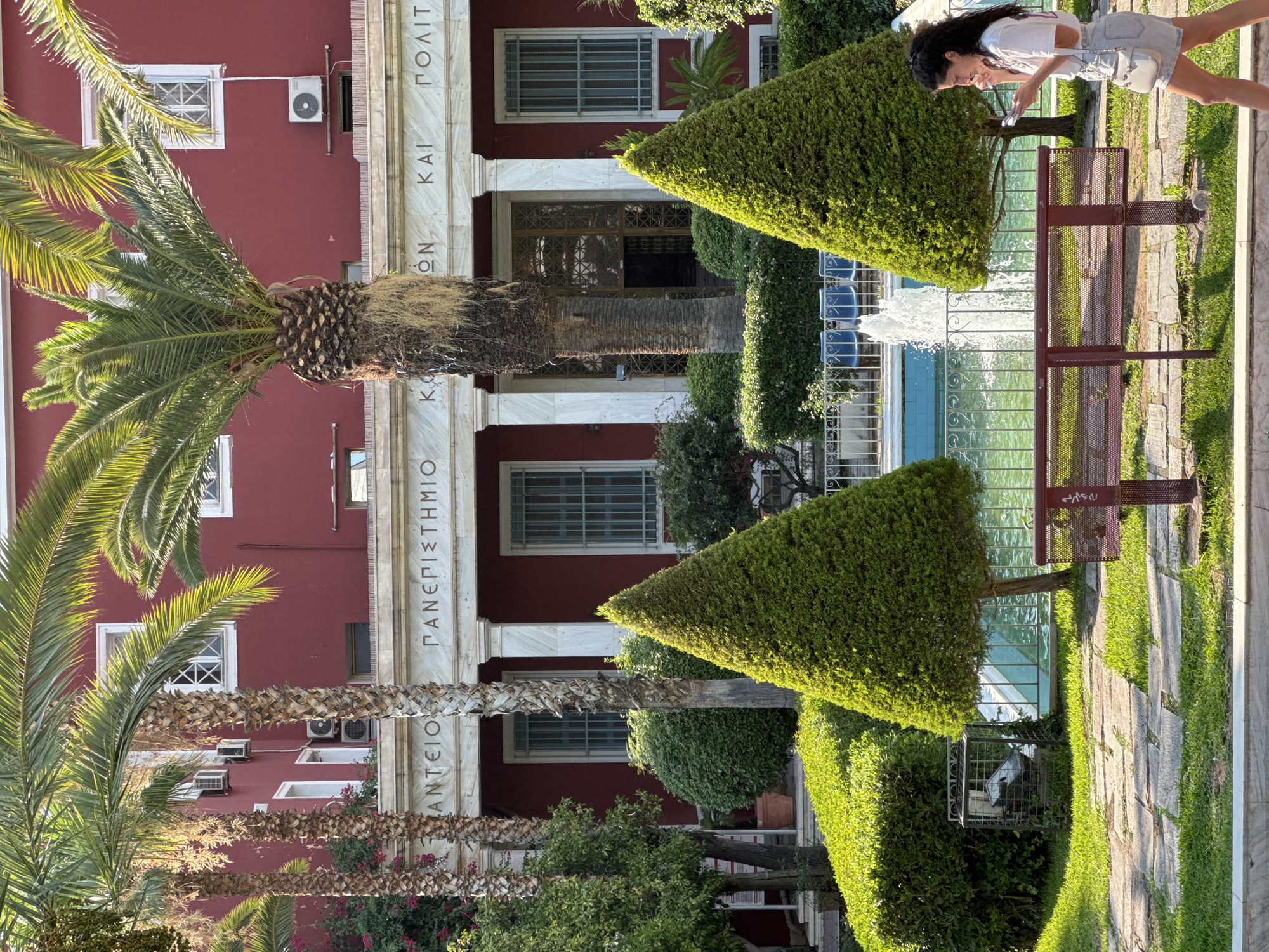 The image captures a sunny outdoor scene in front of a red building with white columns and marble inscriptions. Palm trees and manicured greenery, including conical topiaries, dominate the foreground. A fountain sprays water in a pool behind a decorative fence. A woman with dark curly hair, wearing a white t-shirt and denim shorts, walks past a red bench, looking at her phone. The building appears to be a public institution, possibly an educational or administrative building, given the inscriptions and architecture. The overall impression is one of a well-maintained public space, likely in a Mediterranean climate, consistent with Athens, Greece.