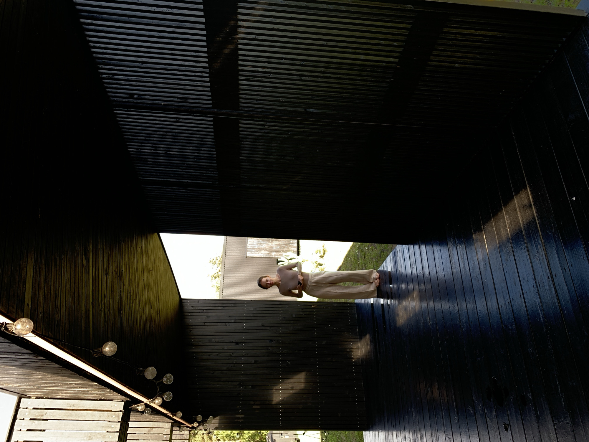 A medium shot shows a person standing on a dark, glossy wooden deck. The person, with light brown hair, wears a light brown long-sleeved shirt and loose-fitting beige pants. They are barefoot and holding a dark object in their right hand. Behind them, a black slatted wooden wall extends, with a large opening to the right revealing another similar structure in the background, which appears to be part of a building or pavilion. The deck is lit by sunlight filtering through string lights hanging from the dark wooden ceiling of the structure. The environment suggests an outdoor, possibly rural or suburban, setting with glimpses of greenery and a bright sky visible through the openings. The overall scene is characterized by strong contrasts between light and shadow due to the direct sunlight.