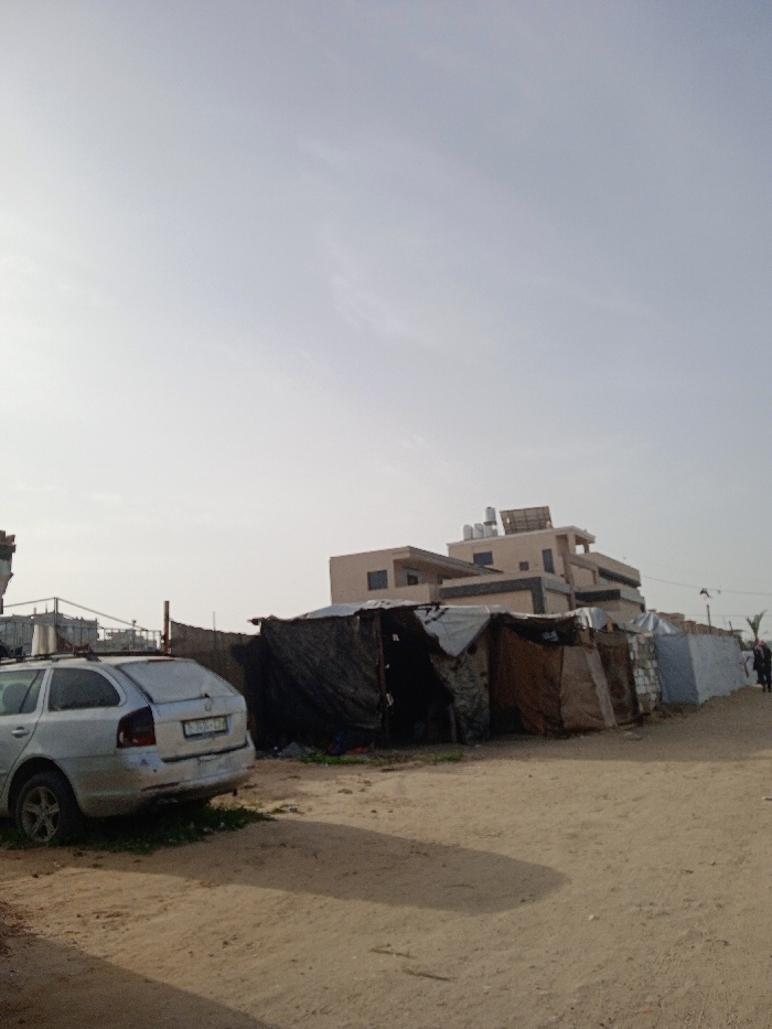 This image depicts a dusty, unpaved urban landscape in Khan Yunis, Palestinian Territory. In the foreground on the left, a silver station wagon, possibly a Skoda Octavia model, is parked facing away from the viewer, angled slightly left. Its rear bumper appears damaged, and a Palestinian license plate (green characters on white) is visible but partially obscured. The vehicle rests partially on a small patch of green vegetation on the dirt ground.
In the midground, running from the center-left towards the right, is a row of approximately five to seven makeshift shelters. These structures are constructed from various salvaged materials including dark tarpaulins (black, grey, brown), other fabrics, and possibly corrugated metal. They exhibit irregular shapes and appear rudimentary. Debris is scattered on the ground directly in front of these shelters.
Behind the makeshift shelters, a multi-story, light-colored (beige/tan) building stands, featuring multiple windows and a flat roof equipped with what appear to be solar water heaters or panels. A fence separates the row of shelters from this more permanent structure. To the far right, two indistinct figures are visible in the distance, walking away from the viewer on the dirt path. The sky is a pale, hazy blue-grey with minimal cloud cover, and long shadows are cast across the unpaved ground, suggesting morning or late afternoon light.