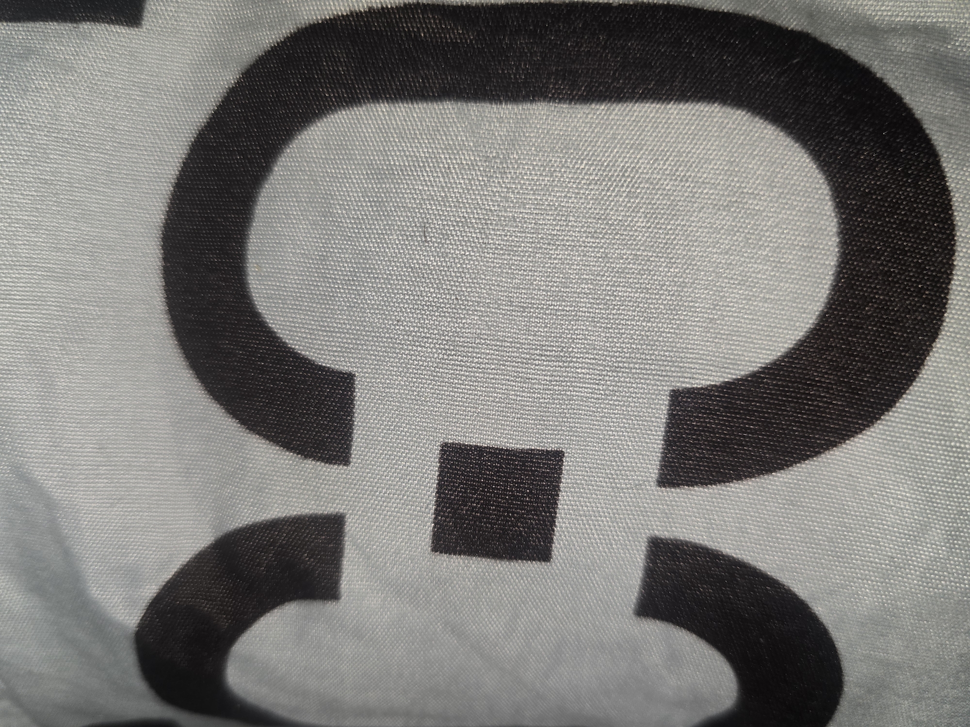 The image displays a close-up, abstract view of a black symbol on a light-colored, textured fabric. The symbol appears to be a stylized representation of the number "8" or an infinity symbol, formed by two large, rounded shapes connected by a smaller square in the center. The fabric has a visible weave pattern, suggesting it might be a piece of clothing, upholstery, or a banner. The lighting creates subtle shadows and highlights on the fabric, emphasizing its texture. The image does not contain any information that would allow for its localization in Ulan-Ude, Russia.