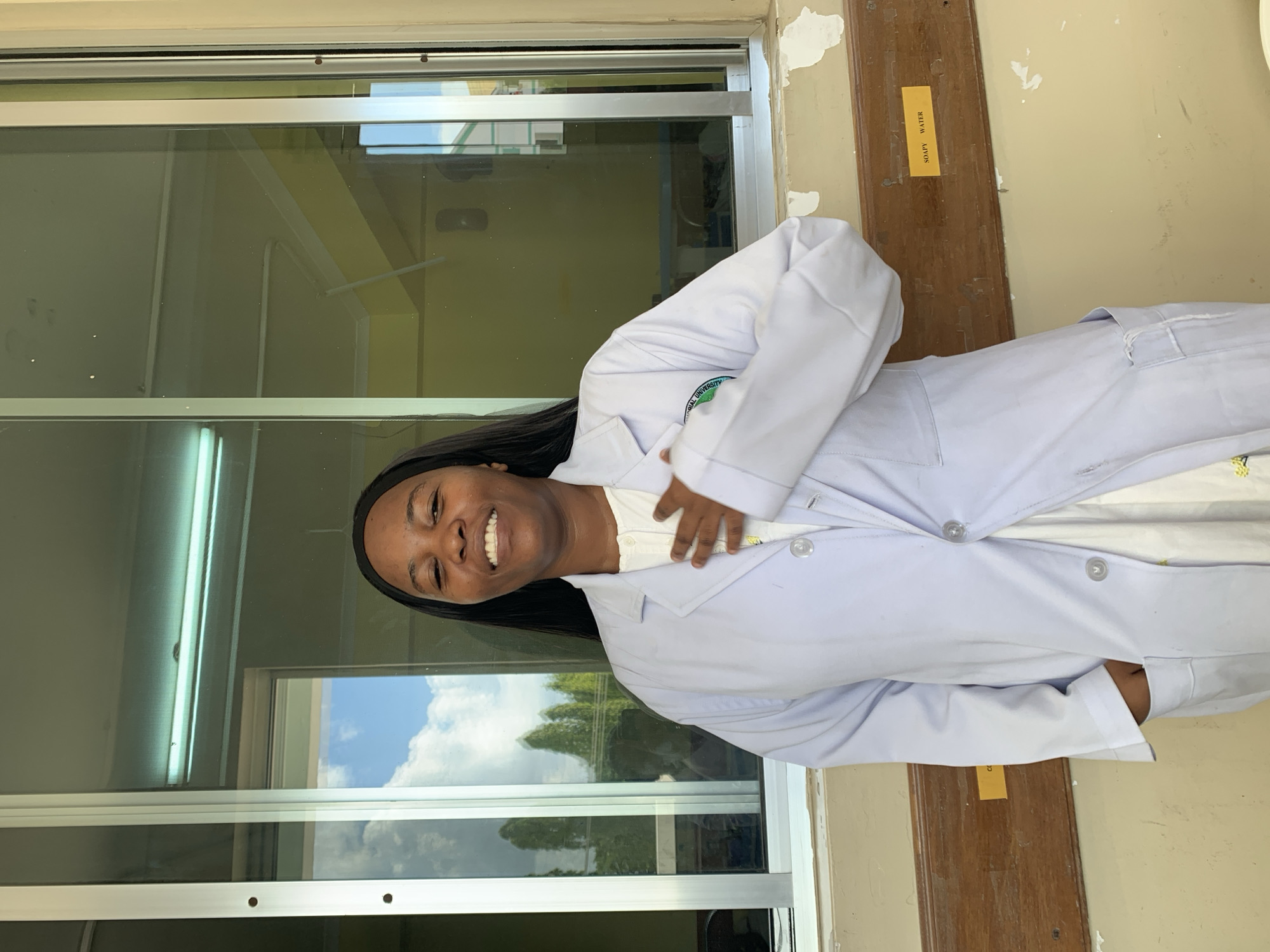 A young woman wearing a white lab coat is smiling and standing indoors, likely in a laboratory or medical facility, based on her attire. Reflected in the window behind her are trees and a blue sky with clouds, suggesting an outdoor setting. A sign that reads "Soapy Water" is visible on the wall. While I cannot confirm the location with certainty based on the image, and I am unable to infer that she is in Dar es Salaam, Tanzania.
