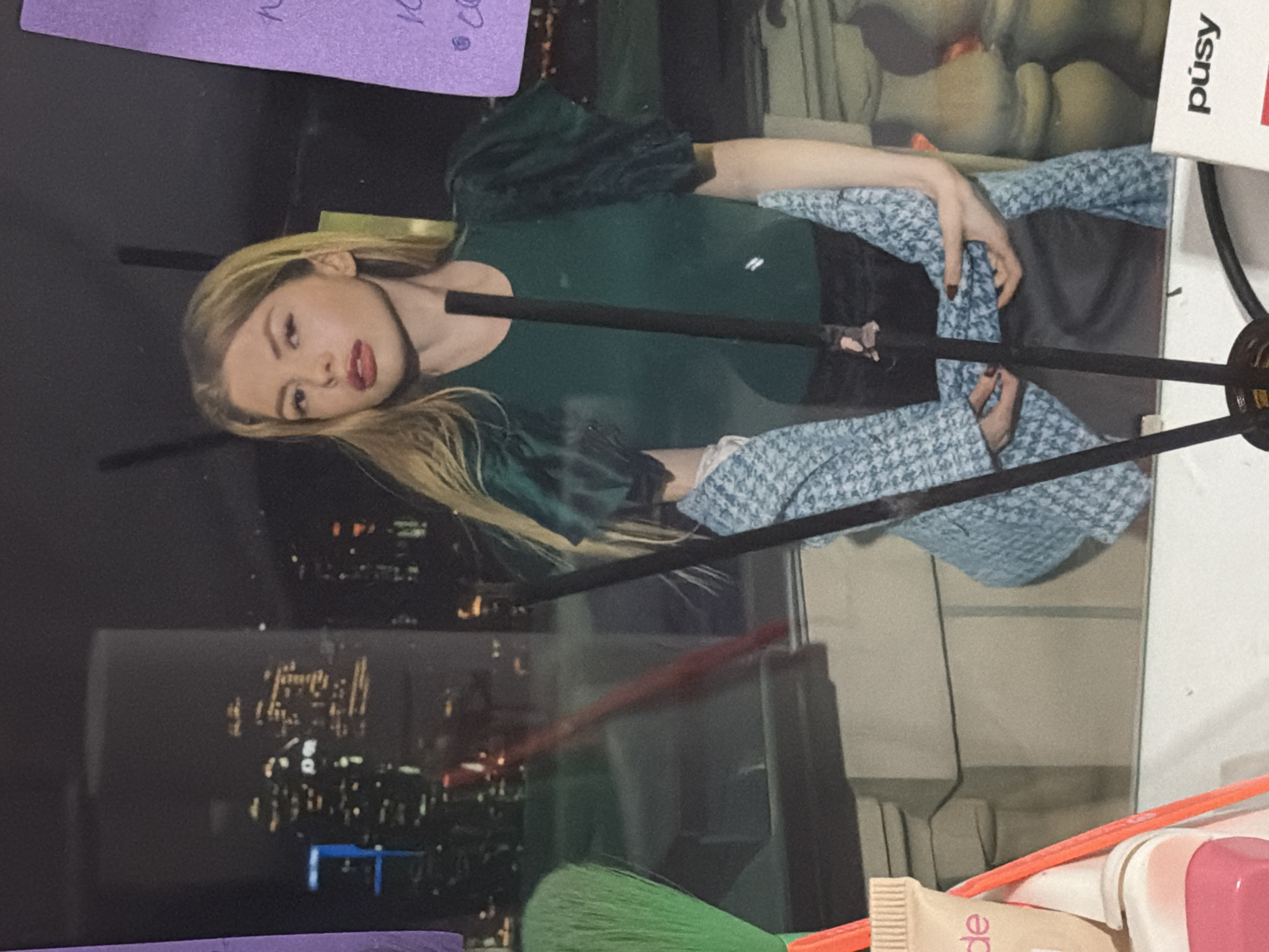 A blond woman is reflected in a mirror. She is wearing a dark green, puffed-sleeve shirt, a patterned blue jacket, and a black skirt. Her hair is long and blonde, and she is wearing dark lipstick. In the background of the reflection, a city skyline at night can be seen, with buildings and lights indicating it is likely Moscow, Russia. On the left side of the image, a green makeup brush and a tube of makeup are visible. On the right, a purple sticky note with handwriting and a white box with the word "púśy" are present. There are also some dark vertical sticks, possibly incense sticks, in the foreground. The overall setting appears to be indoors, possibly a room with a view of the city.