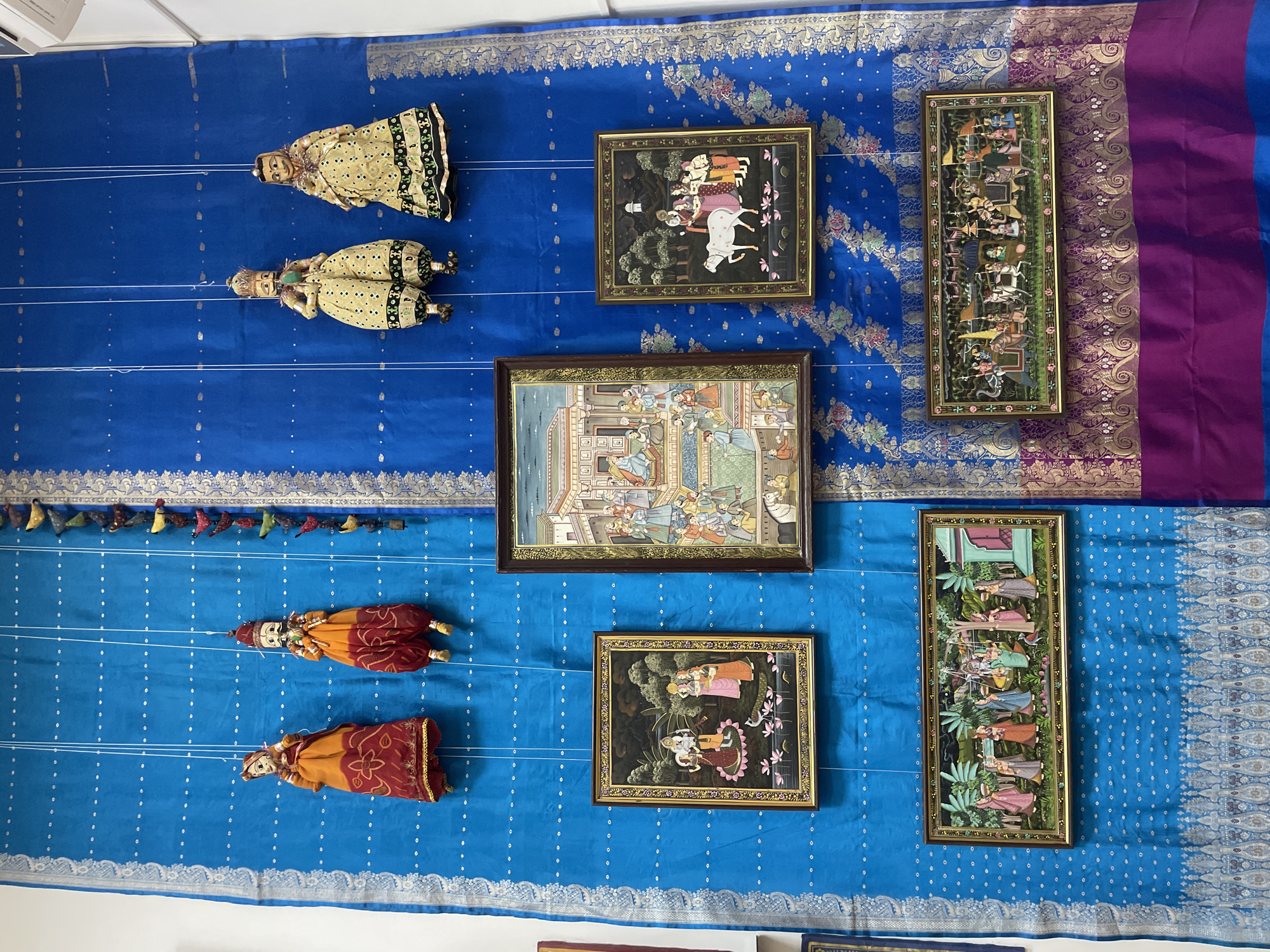 The image displays a vibrant collection of traditional Indian artifacts, arranged as a decorative or exhibition display against a blue textile background.
The subjects include four traditional Indian string puppets, known as Kathputli. The upper pair, positioned on the left, are dressed in light green/yellow and black garments. The lower pair, also on the left, are attired in orange and red. All puppets are suspended by thin white strings. Above the lower puppet pair, a string of small, colorful fabric tassels hangs.
Interspersed among the puppets are five framed miniature paintings. The top-left painting depicts multiple figures and a white cow in a landscape. Below it, a larger, horizontally oriented painting showcases an elaborate indoor court scene with numerous figures. On the right side of the display, two long, vertically oriented paintings feature various scenes with human figures, horses, and elements of architecture and nature. A fifth, smaller painting in the bottom-middle section illustrates two pairs of figures, possibly Radha and Krishna, in a garden setting with trees and flowers.
The background consists primarily of a rich blue fabric, likely a sari or decorative cloth, adorned with intricate gold and silver woven patterns along its borders and scattered silver dots throughout. A vertical section of purple fabric with gold patterns is visible on the far right, providing an accent to two of the paintings.
The activity taking place is the static display of these cultural items. The arrangement is visually engaging, highlighting the detailed artistry of the puppets and paintings. The context indicates this display is located in Sochi, Russia, suggesting an international cultural exhibit. There is no visible text in the image.