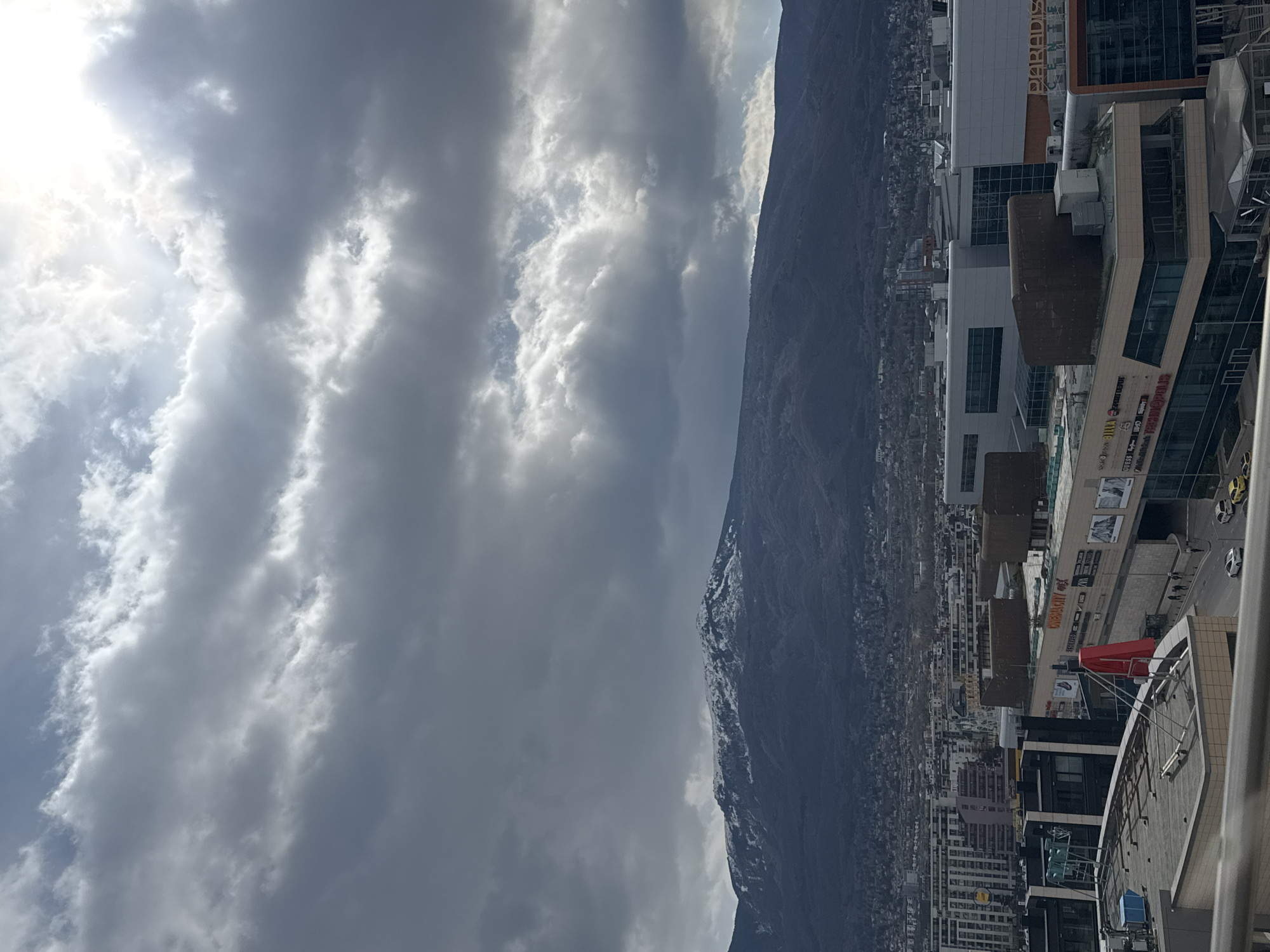 The image shows a cityscape dominated by a large mountain range, likely Vitosha Mountain, with a city sprawling at its base. The foreground features rooftops and buildings of the city, with some commercial complexes clearly visible. The sky is filled with heavy clouds, suggesting overcast weather. The city appears to be Sofia, Bulgaria, given the distinct presence of Vitosha Mountain.