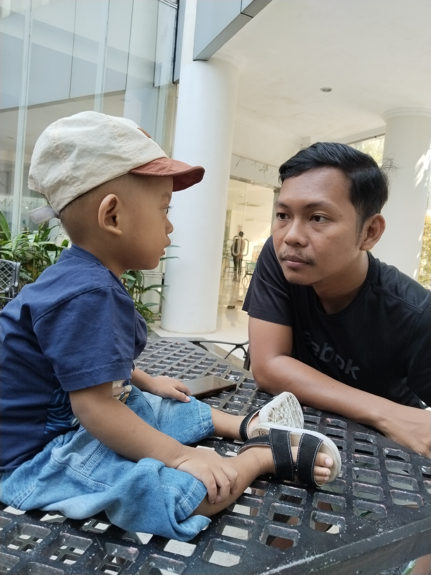 A medium shot captures two males, an adult and a young child, seated at a black metal mesh table outdoors. The adult, positioned on the right, wears a black t-shirt and has short dark hair and a mustache, looking towards the child. The child, on the left, wears a blue t-shirt and denim shorts, with a light-colored cap with a brown brim, and is looking at the adult. Both are seated. The environment suggests an urban setting, possibly a cafe or public seating area, with a building's facade and columns visible in the background, along with some greenery. The lighting is natural, indicating daytime.