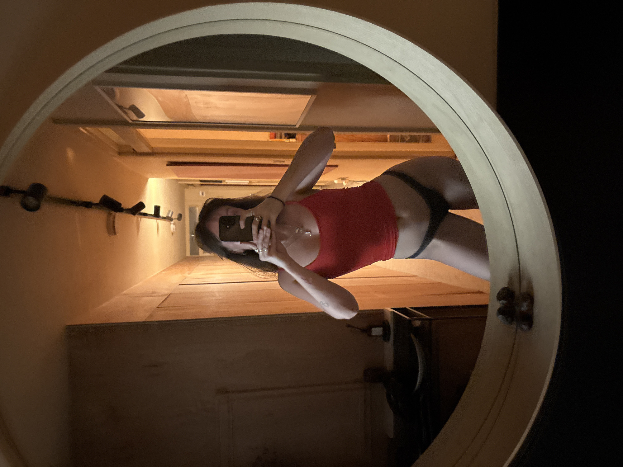 A person is seen in a mirror selfie, captured within a large, circular, wall-mounted mirror with a light-colored frame. The person, appearing female with long dark hair, is wearing a red top and dark-colored underwear or bottoms. She holds a smartphone to take the picture, partially obscuring her face. A necklace with a pendant is visible around her neck, and a bracelet is on one wrist.
The reflection in the mirror reveals an indoor setting, likely a narrow hallway or room, with warm, dim lighting suggesting evening or night. The walls appear light-colored, possibly beige or cream. A wall-mounted light fixture with spotlights is reflected on the left side, and a rectangular feature, possibly a vent or framed item, is seen above the person's head in the reflection. Below the reflection of the person, a wooden surface or low platform is visible. The internal architecture suggests a residential or hotel environment in London, United Kingdom. No discernible text is visible in the image.