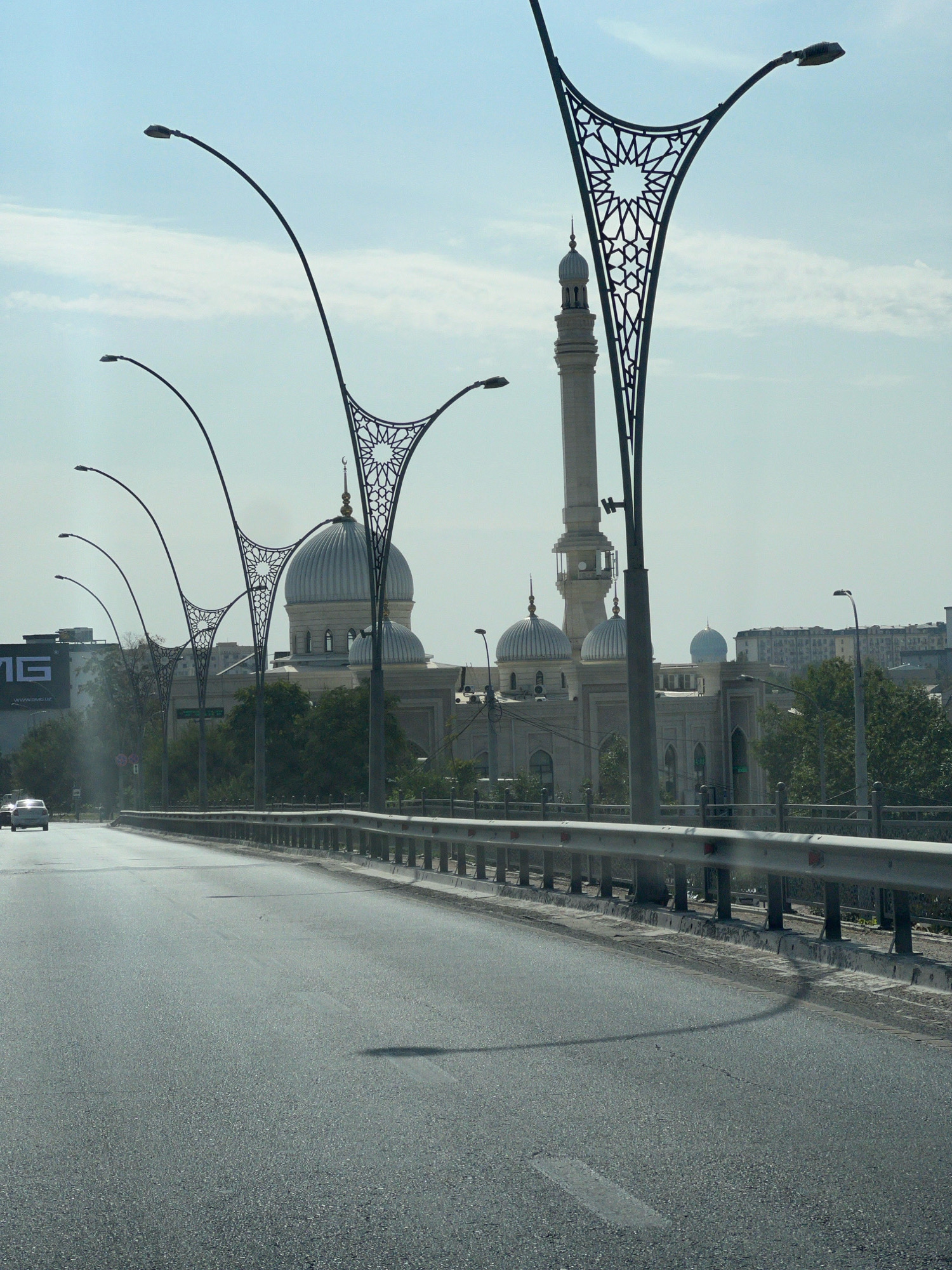 This image captures a view of a modern mosque in Tashkent, Uzbekistan, seen from a road that appears to be a bridge or overpass. The foreground is dominated by the asphalt road, with a metal guardrail lining the right side. Several cars are visible in the distance on the left side of the road. The background features the mosque, characterized by its prominent minaret and multiple domes, one of which is a striking light blue. Ornate streetlights with Islamic geometric patterns are strategically placed along the road, framing the view of the mosque. The sky is a clear, pale blue with faint clouds. The overall scene suggests a sunny day in an urban environment.