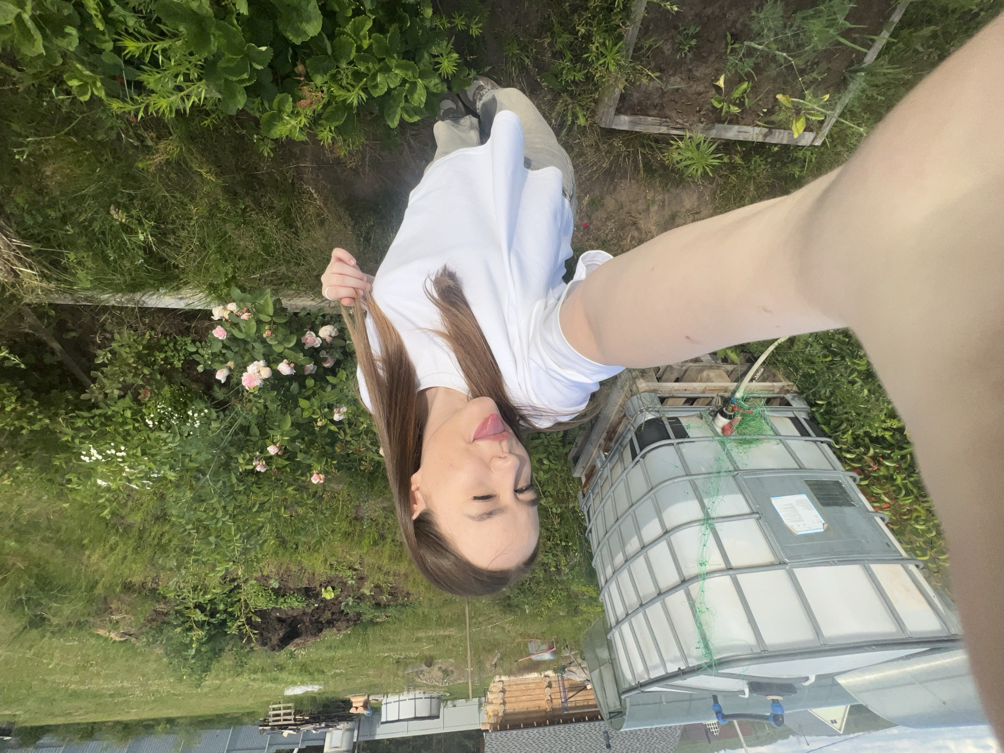 A young woman with long brown hair is taking a selfie in an outdoor, rural setting. She is wearing a white t-shirt and light-colored pants. Her facial expression is playful, with one eye winked and her tongue sticking out. She is standing in front of a large white IBC tote tank, which appears to be used for water storage. In the background, there is a wooden structure that resembles a small house or shed, a garden with green plants and some pink roses, and a metal fence. The overall impression is that of a garden or yard area in a semi-rural or dacha environment, possibly in the city of Chaikovsky, Russia, given the typical appearance of such structures and vegetation. The lighting suggests it is daytime, with the sun casting shadows.