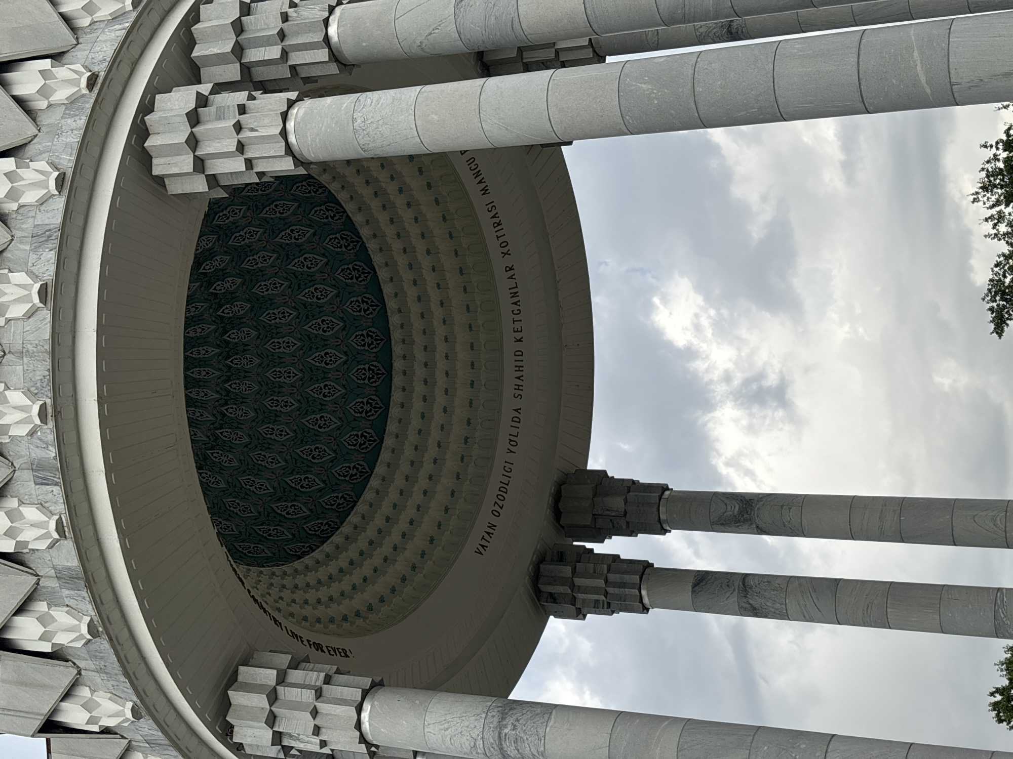 The image shows a detail of the Monument of Independence in Tashkent, Uzbekistan. The monument features a semicircular structure with ornate green tiling on the ceiling and Arabic inscriptions around the perimeter. Four large, fluted marble columns support the structure. The sky is overcast with clouds. The inscription reads, "Vatan ozodligi yólida shahid ketganlar xotirasi mangu".