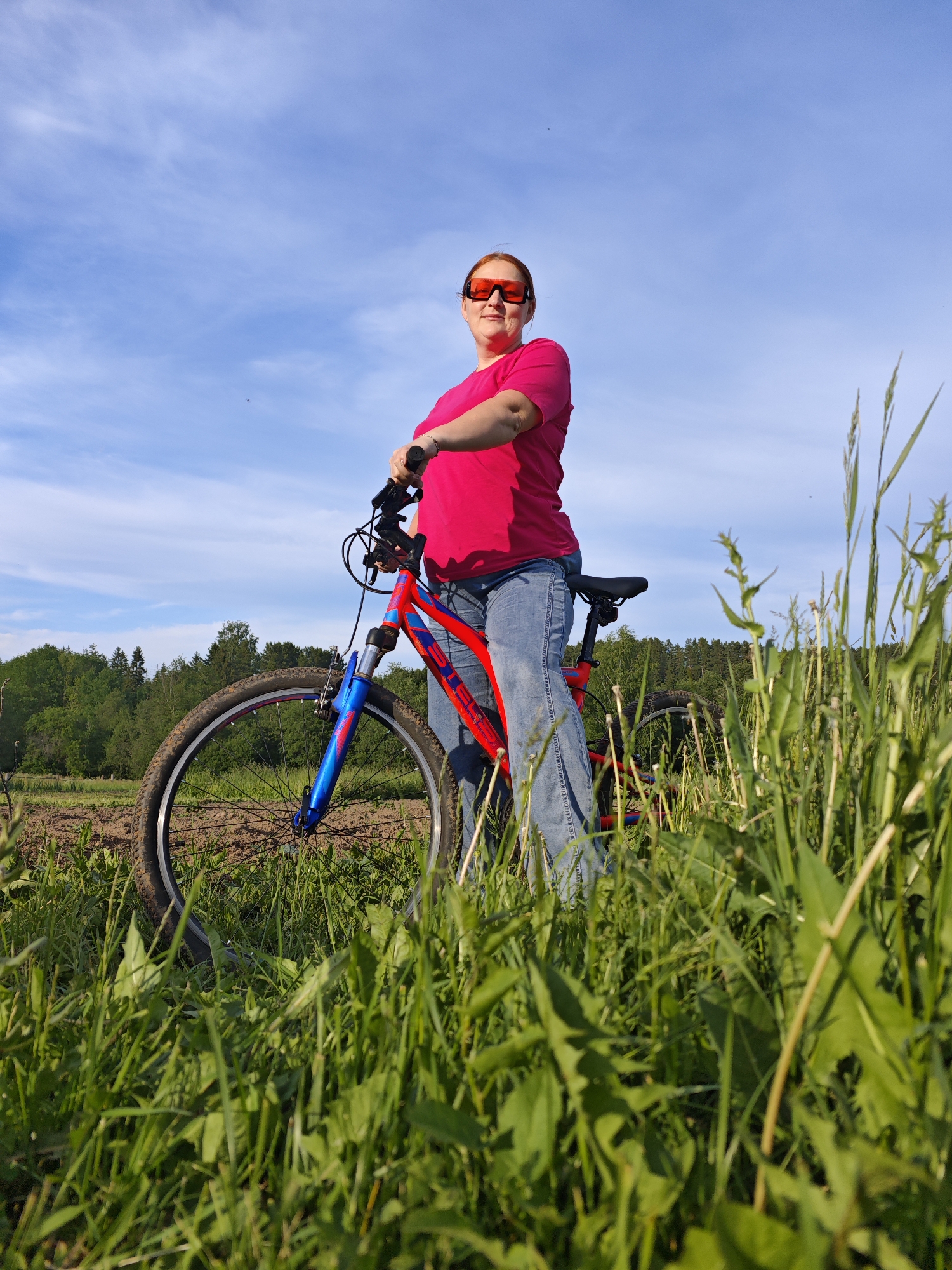 The image captures an outdoor scene featuring an adult woman standing with a mountain bike in a grassy field under a bright blue sky, set in Фоки, Russia. The photograph is taken from a low-angle perspective, emphasizing the subject against the expansive sky.
The main subject is a woman, likely in her 30s or 40s, with reddish hair. She is dressed in a bright hot pink short-sleeved t-shirt, light blue denim jeans, and distinctive bright orange-red rectangular sunglasses. She stands holding the handlebars of her bicycle, looking towards the camera with a calm expression. Her posture suggests she is either resting or posing during a bike ride.
The bicycle is a mountain bike, notable for its striking red frame with blue accents on the front suspension forks. Part of the brand name "STELS" is visible on the bike's downtube. The bike's wheels are partially obscured by lush, tall green grass that dominates the immediate foreground, indicating a natural, unmanicured environment.
In the midground, beyond the grass, there appears to be a section of tilled soil or a dirt path, leading towards a dense line of green trees forming a forest in the background. The sky is clear with a few scattered white clouds, suggesting sunny and pleasant weather conditions, likely late afternoon given the bright, direct light. The overall setting conveys a recreational outdoor activity in a peaceful, rural landscape. No other individuals or significant structures are visible.