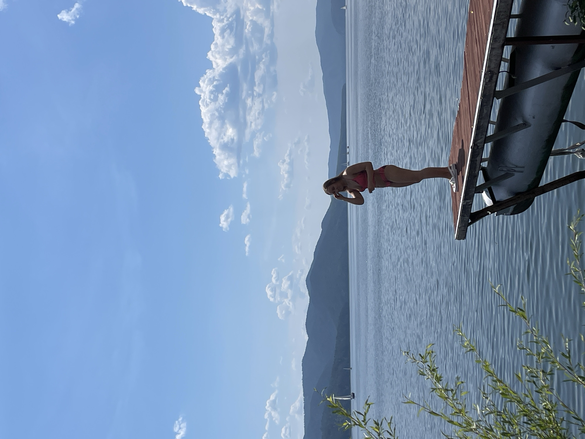 A young woman in a pink bikini stands on a wooden dock on a body of water, with mountains in the background. She is facing to the left, with her right hand touching her hair. The water is blue and calm, with gentle ripples. There are a few sailboats in the distance. The sky is blue with scattered white clouds. The scene appears to be a summer day in a rural or mountainous area. The location is likely near Divnogorsk, Russia, given the mountainous landscape and prominent reservoir.