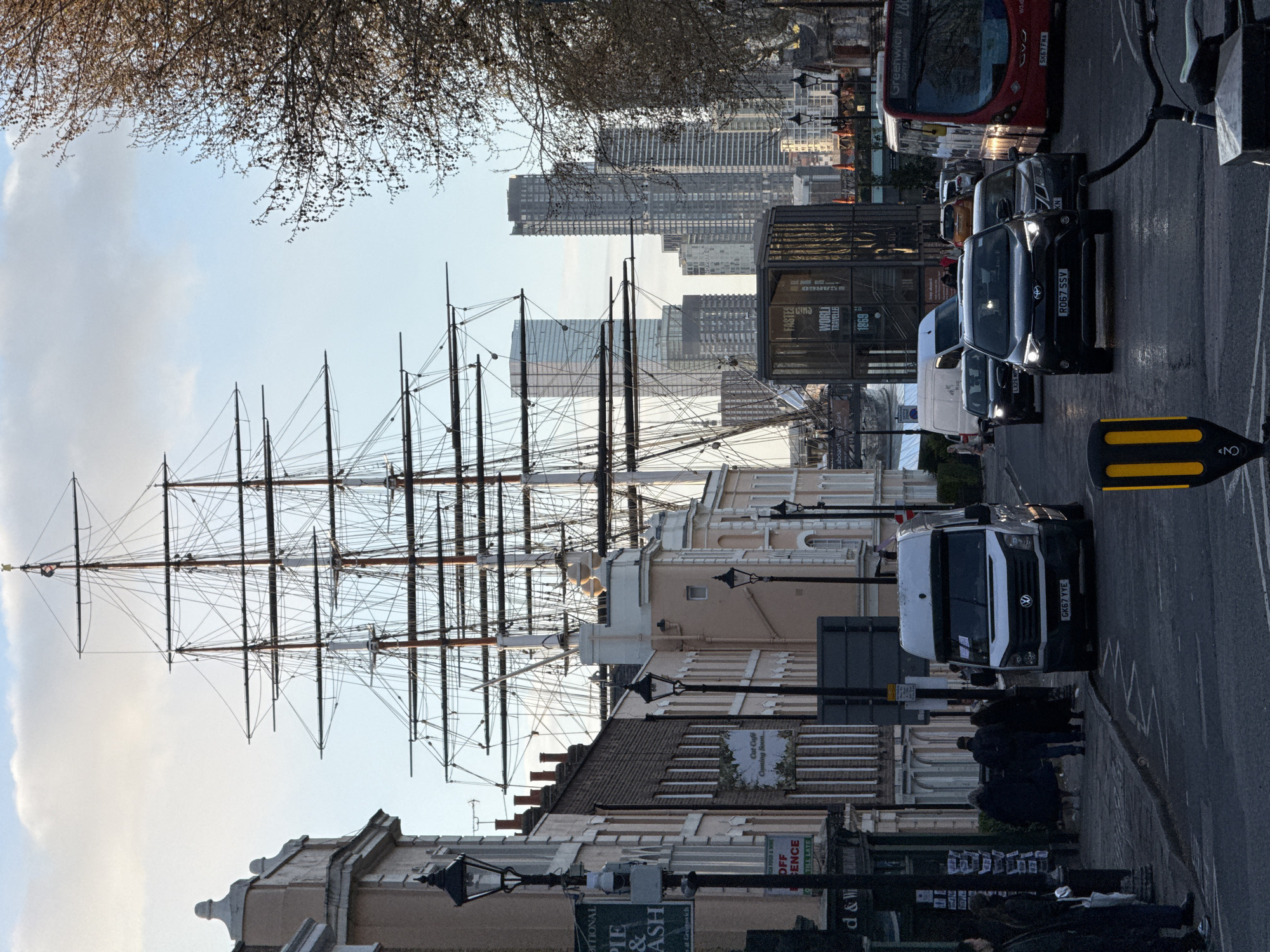 The image shows a street in Greenwich, London, UK, with the Cutty Sark clipper ship's masts visible behind buildings. A row of buildings lines the left side of the street, with shops at street level. Cars and a red double-decker bus are moving or stopped on the right side of the street. Pedestrians are on the sidewalk in front of the shops. Modern skyscrapers are visible in the distance beyond the Cutty Sark.