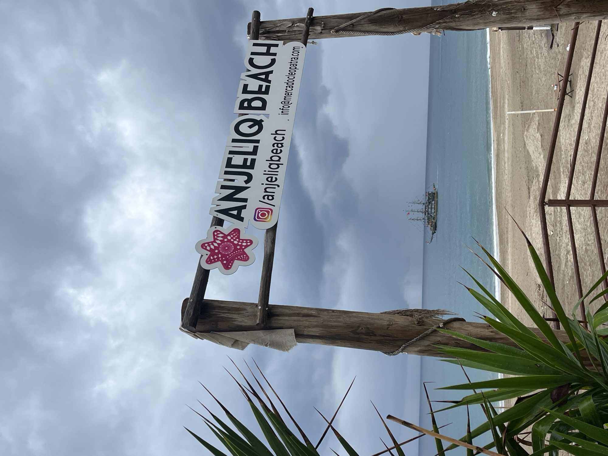The image shows a sign for "Anjelio Beach," located in Alanya, Turkey. The sign is made of wood with white text. The sign lists the website and Instagram handle "/anjeliobeach" and the email address "info@mercadocleopatra.com". A pink flower design adorns the left side of the sign. Behind the sign, the ocean and a beach are visible. A ship with sails is visible in the water. The sky is overcast. A green plant with long, pointed leaves is in the foreground.