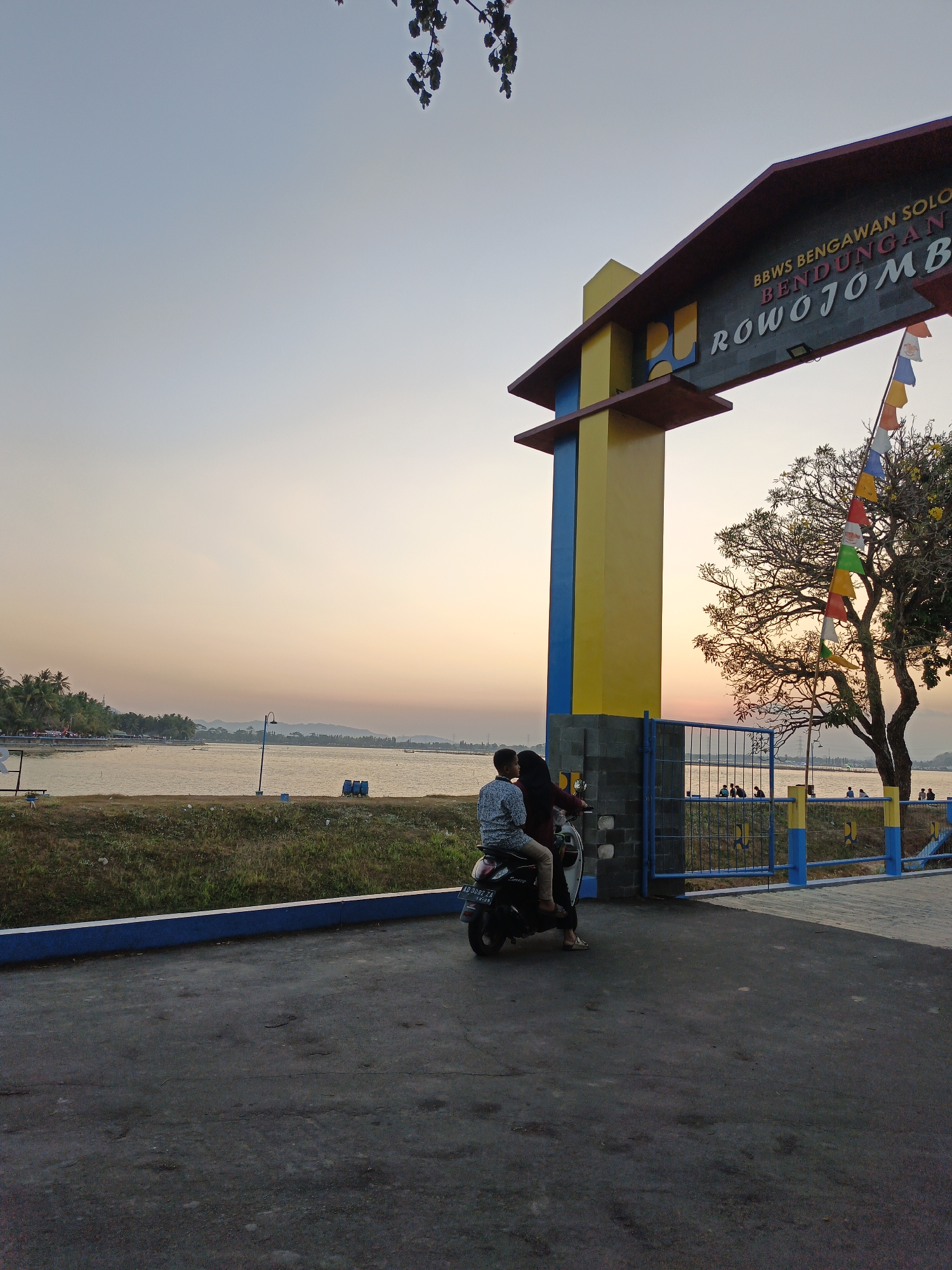 A motorbike is parked in front of a gate at sunset at Rowo Jombor Reservoir, Krakitan, Indonesia. Two people are seated on the motorbike. The gate has a sign that reads "BBWS BENGAWAN SOLO BENDUNGAN ROWOJOMBOR". A tree with colorful flags is to the right of the gate. The reservoir is visible in the background with mountains in the distance. The sky is a gradient of orange and blue, indicating sunset.
