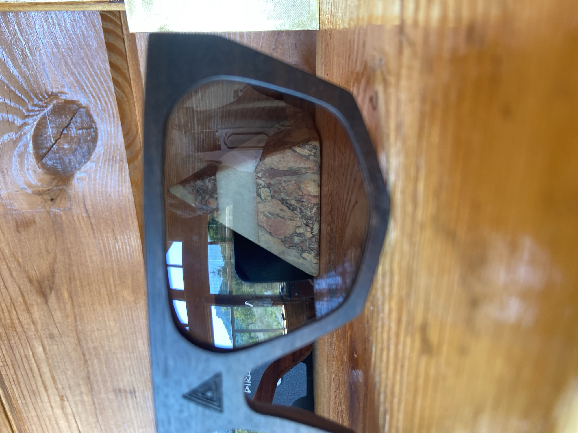 The image shows a close-up of dark-rimmed sunglasses resting on a wooden surface. The lenses of the sunglasses reflect a scene outdoors, with a window showing a landscape of trees and mountains. Inside the reflection, a geometric object, possibly a pyramid made of stone, is visible. The foreground is dominated by the textured wood grain of the surface the sunglasses are on. The background consists of wooden paneling. The image is taken in Красномакское сельское поселение, Ukraine.