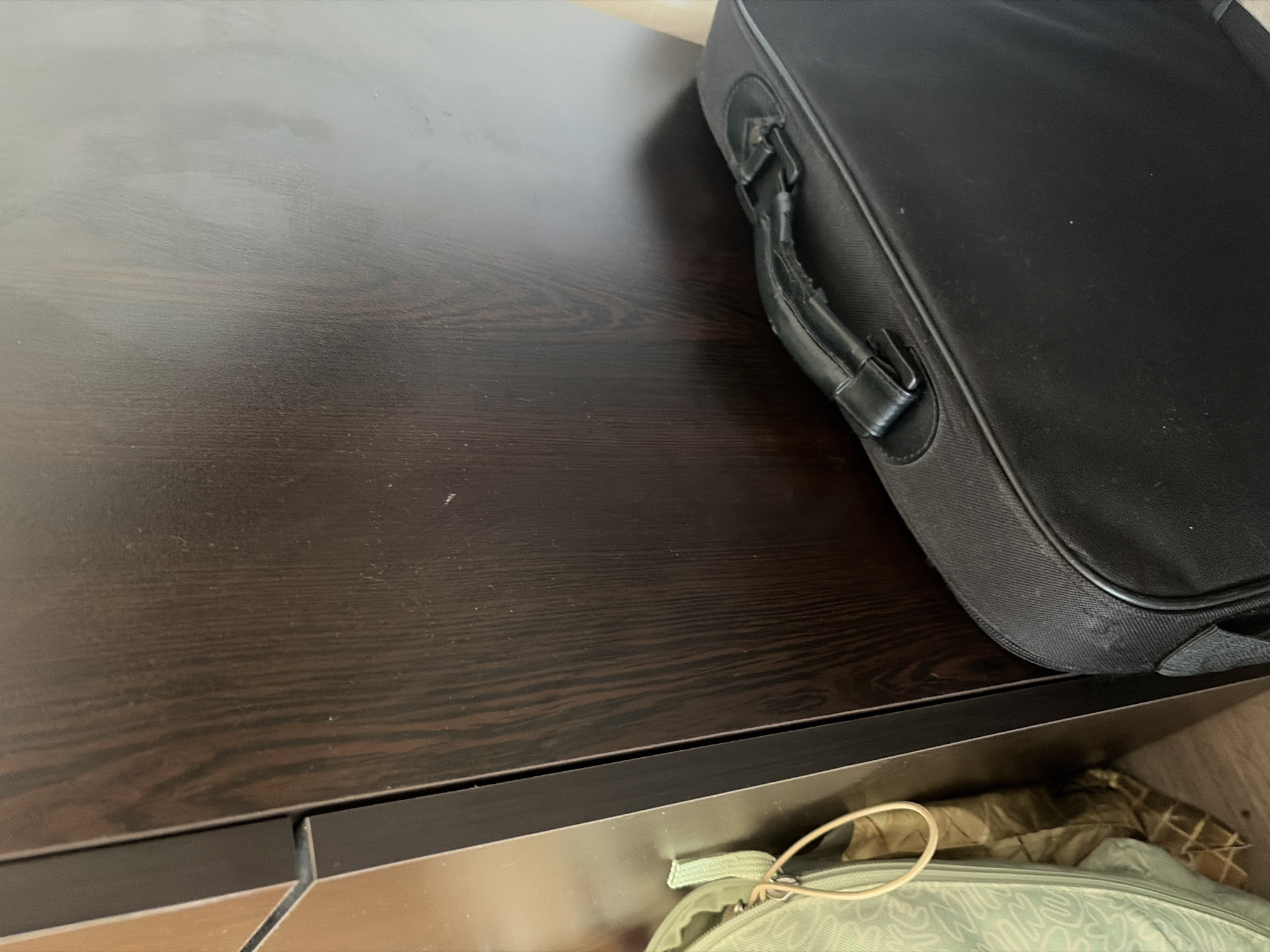 A black rectangular briefcase is placed on a dark wood-grain surface, likely a cabinet or dresser. The briefcase's handle is visible, showing signs of wear. A portion of a light green bag with a drawstring is visible near the edge of the wooden surface, in the lower right corner. The environment suggests an interior setting with no visible indications of location. The city of Sofia, Bulgaria, is not discernible from the image.