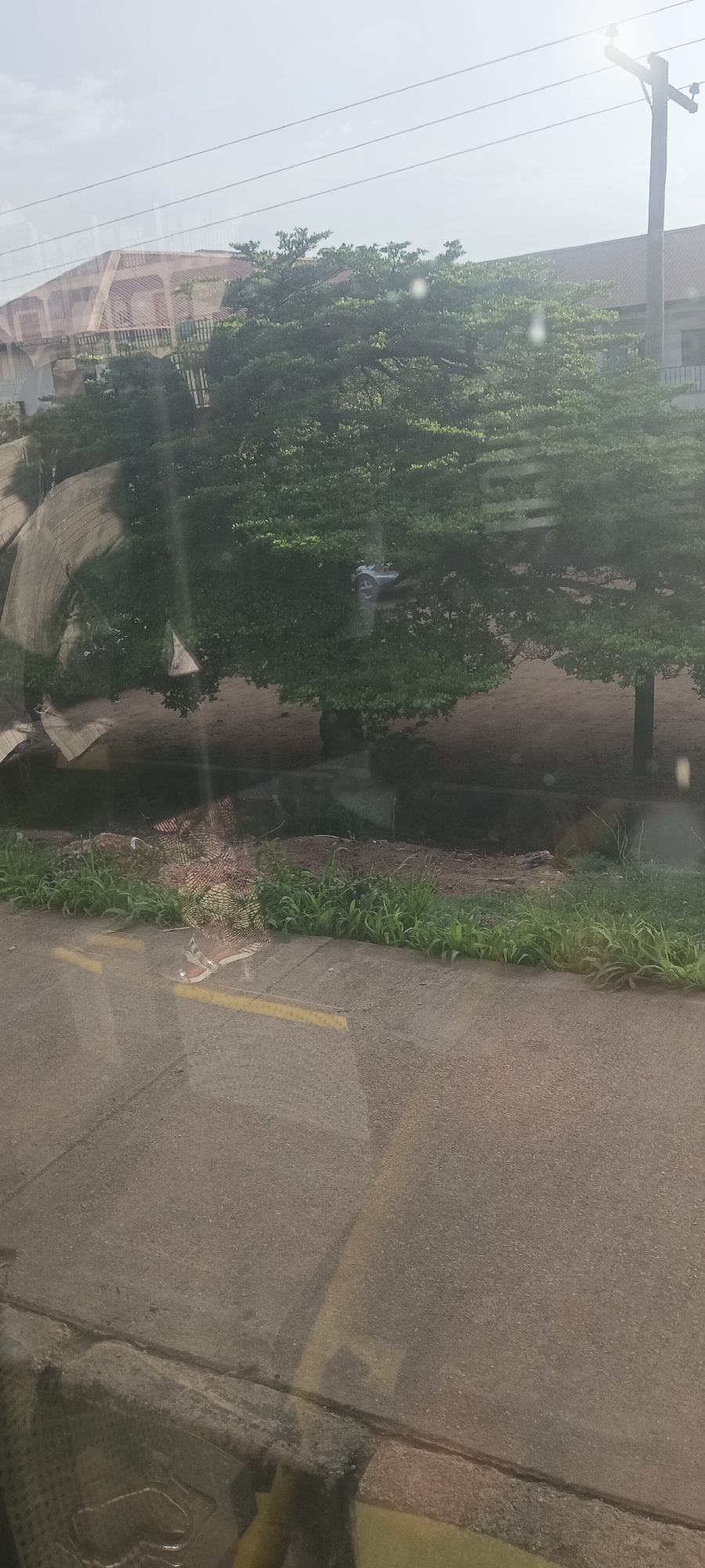 This media file depicts an outdoor scene in Gura Top, Nigeria, viewed through a glass surface, likely a vehicle window, as evidenced by reflections and sun glare. The time of day is bright, likely morning or late afternoon, indicated by a prominent lens flare in the upper right quadrant.
In the foreground, a grey asphalt road or paved area is visible, marked with faint yellow dashed lines. A strip of green grass separates this paved surface from a wide expanse of light brown, bare earth or sand. The midground is dominated by a large, leafy green tree with dense foliage, providing shade to the ground beneath. A white plastic bag or piece of litter is discernible caught within the branches of this tree.
In the background, behind and above the tree, several buildings with reddish-brown tiled roofs and light-colored walls are partially visible. One building features distinct arched window or porch designs. Utility infrastructure includes multiple power lines stretching across a bright, hazy sky, supported by a wooden utility pole on the far right. No people or specific activities are observed. There is no visible text in the image.